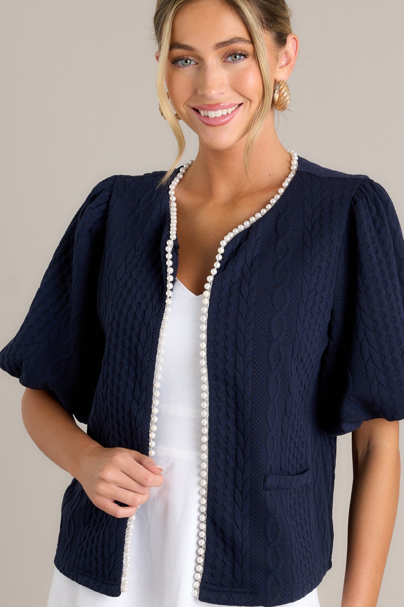 Front angled view of a navy short sleeve jacket featuring a v-neckline, a hook & bar closure, faux pearl trim, a quilted design, faux pockets, and elastic cuffed puff sleeves