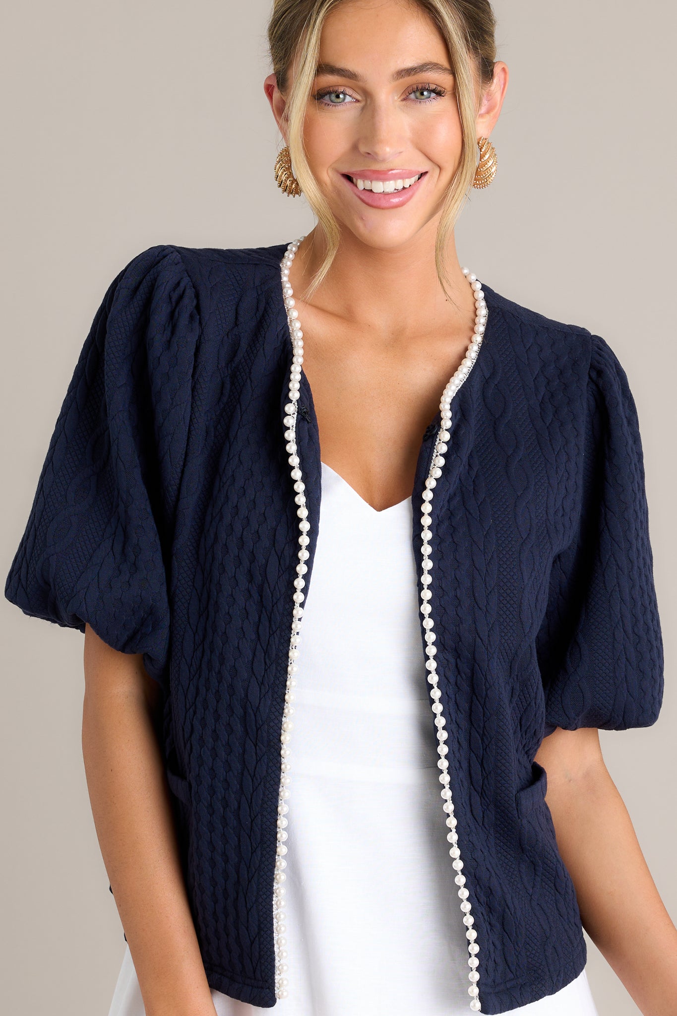 Front view of a navy short sleeve jacket featuring a v-neckline, a hook & bar closure, faux pearl trim, a quilted design, faux pockets, and elastic cuffed puff sleeves.