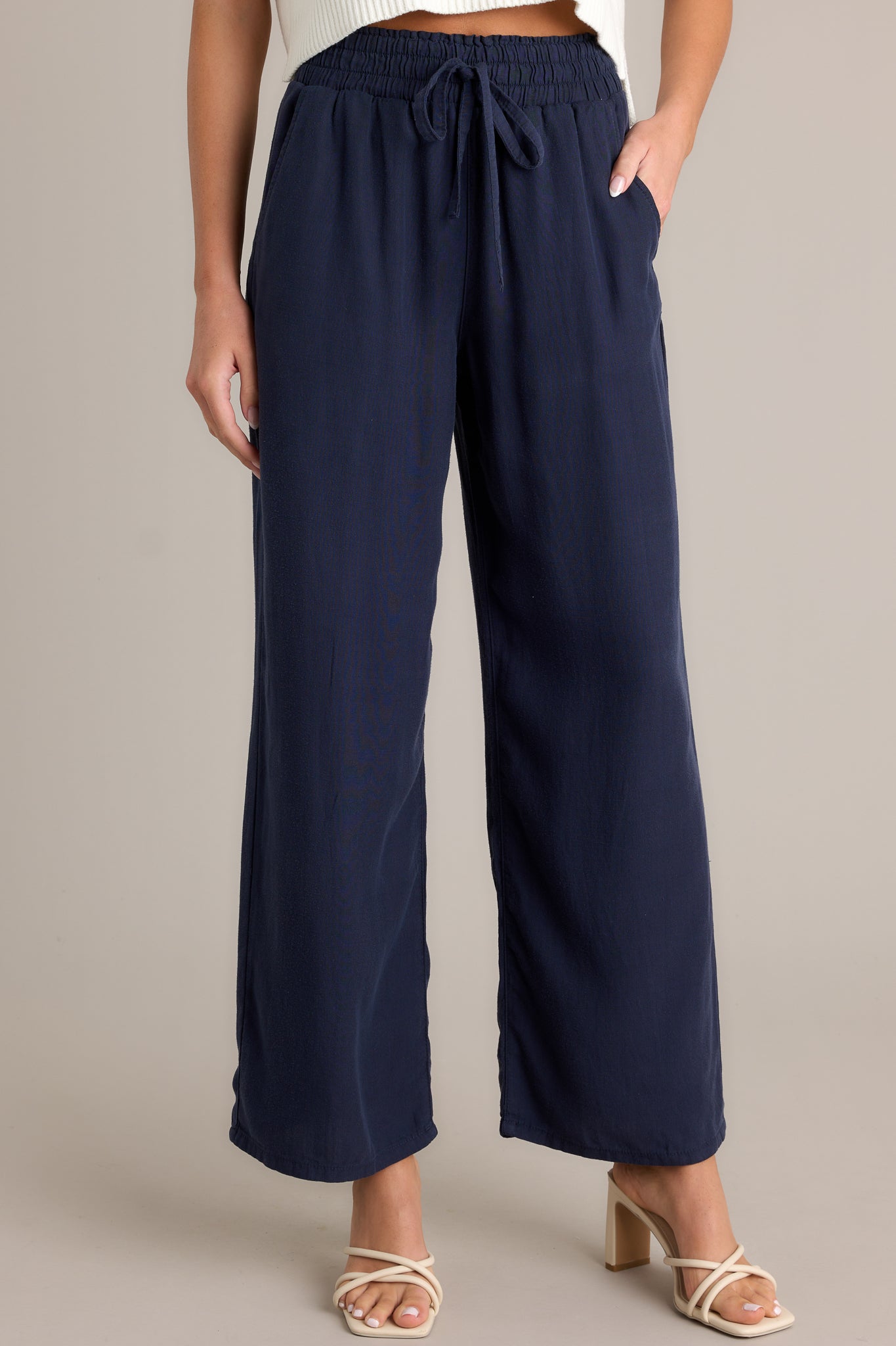 Front view of the material of these navy pants that feature a high waisted design, an elastic waistband with a self-tie drawstring, functional front & back pockets, and a wide leg.