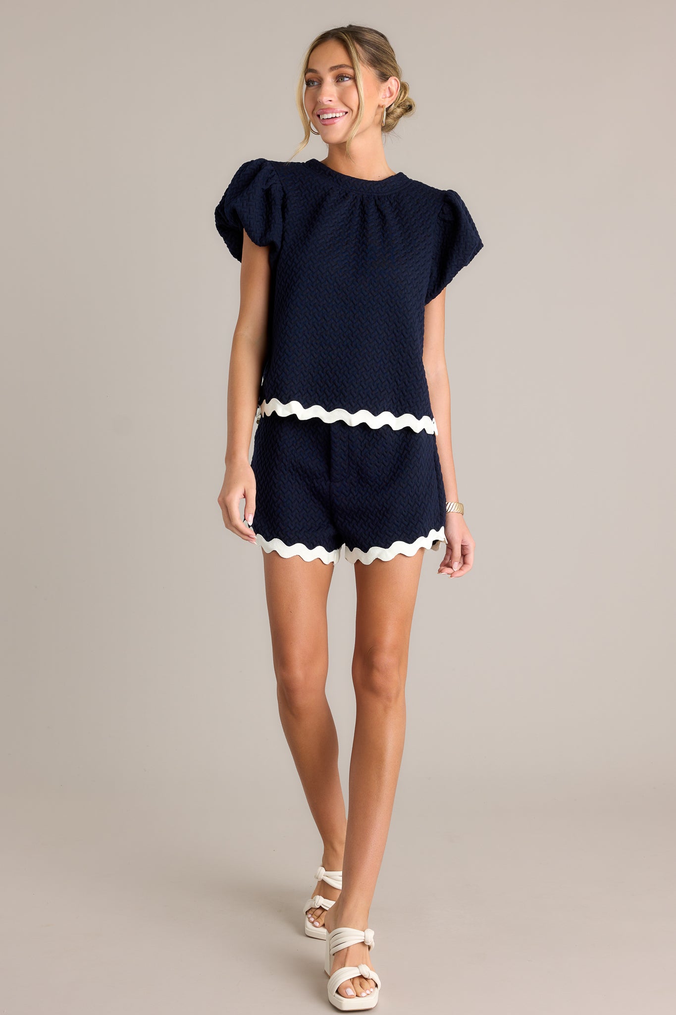 Action shot of a navy top displaying the fit and movement, highlighting the high rounded neckline, quilted material, contrasting scalloped hemline, and elastic cuffed puff sleeves.