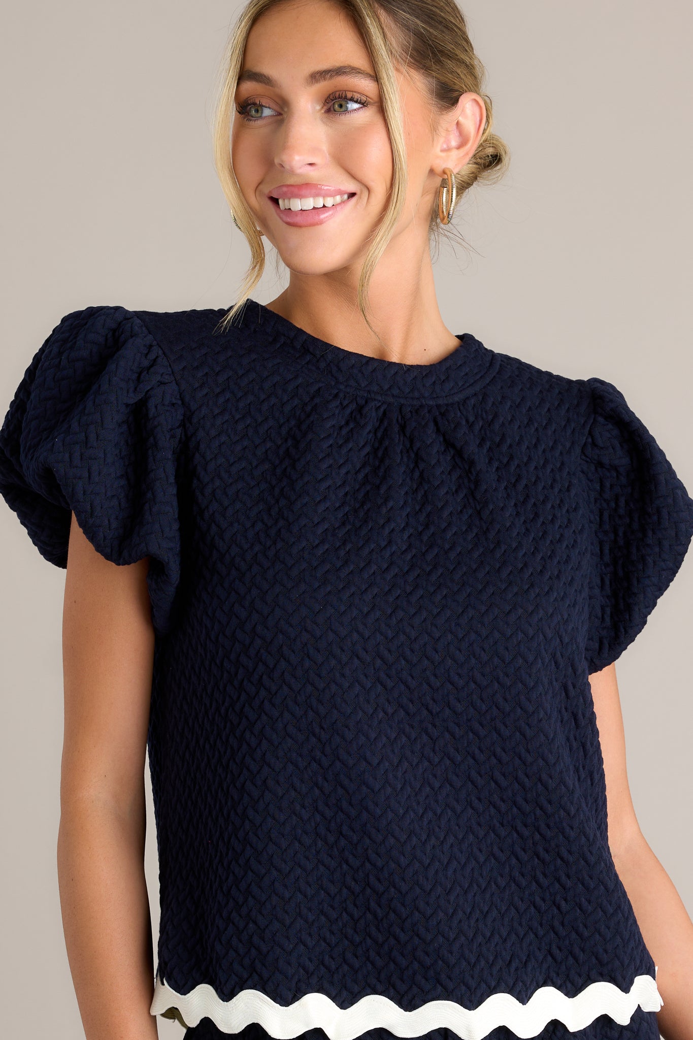 This navy top features a high rounded neckline, a quilted material, a contrasting scalloped hemline, and elastic cuffed puff sleeves.