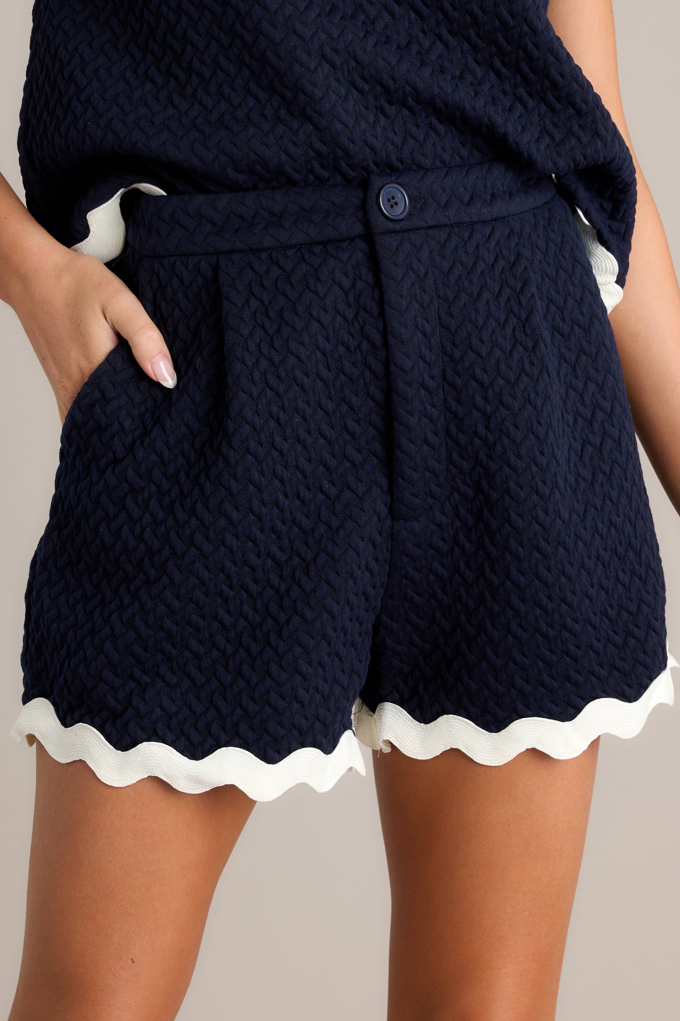 Close-up of the navy scallop shorts showing the zipper and button closure, pleated details, and scallop hemlines.