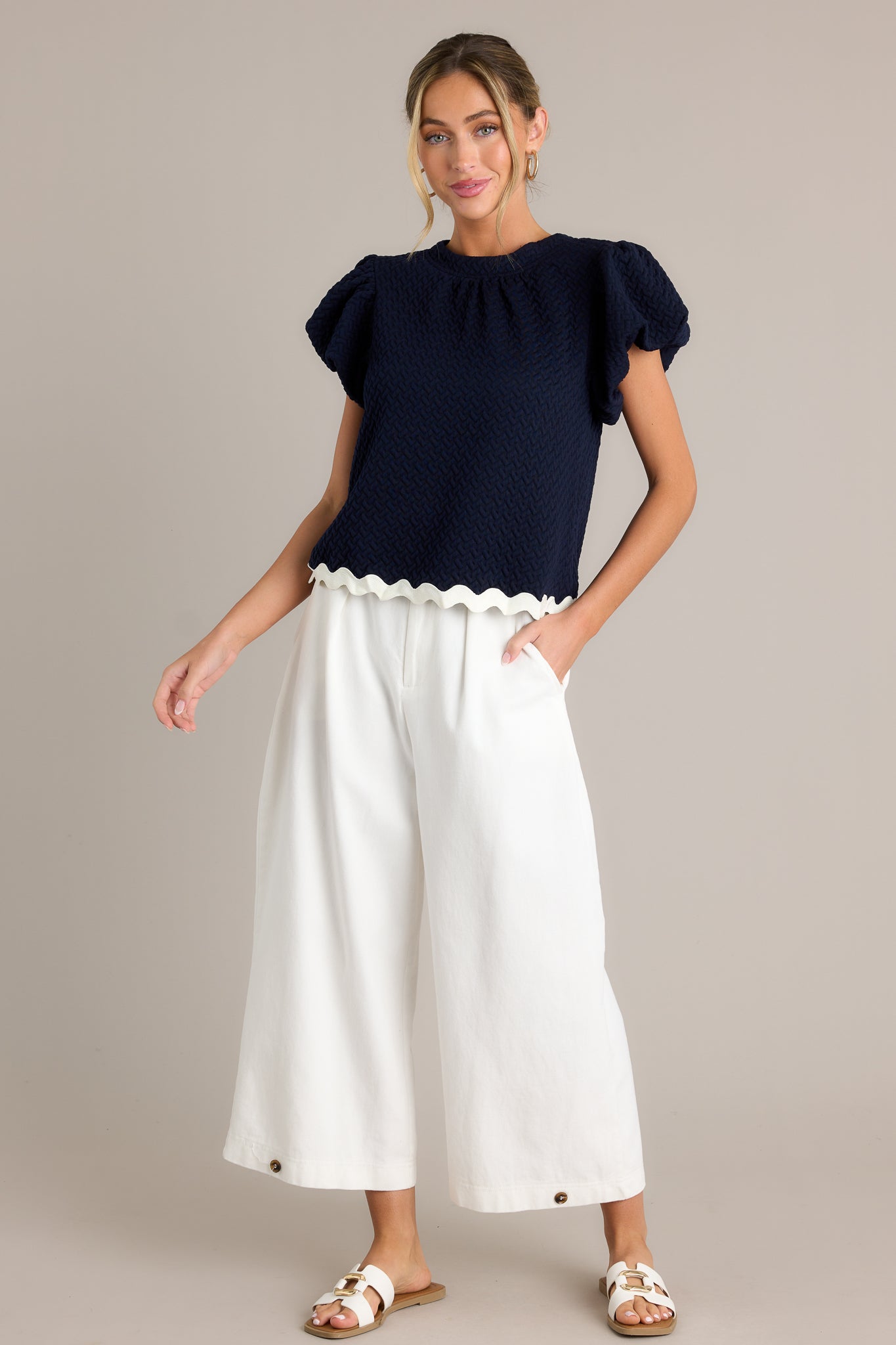 Front angled view of a navy top featuring a high rounded neckline, a quilted material, a contrasting scalloped hemline, and elastic cuffed puff sleeves