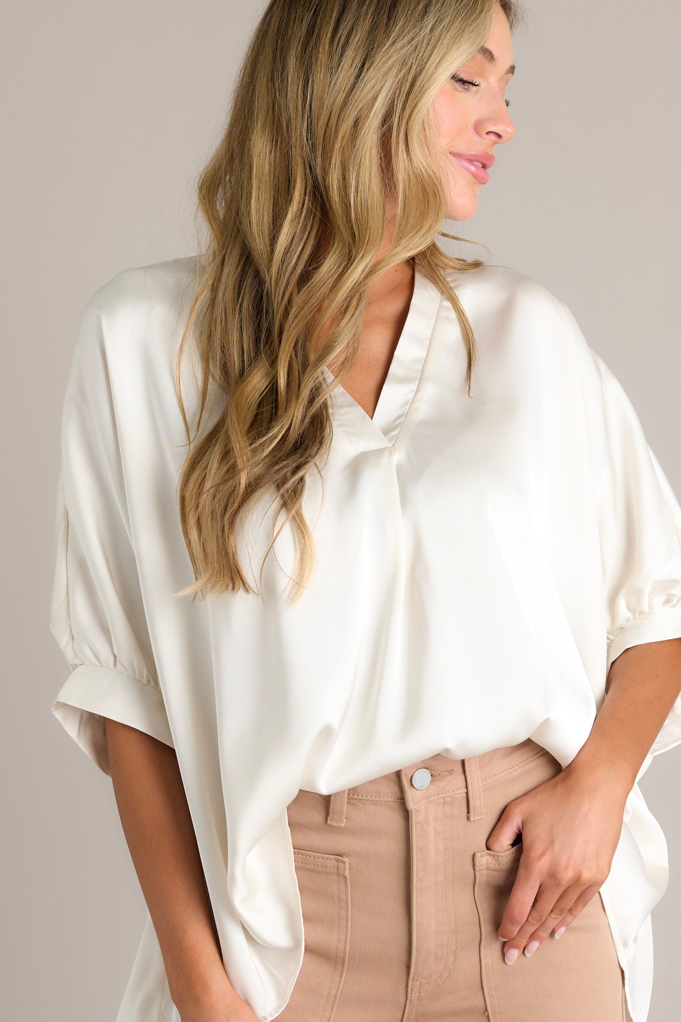 Front angled view of a white top featuring a v-neckline, short dolman sleeves with pleated detailing, a wide, flowy fit, and a satin-like material