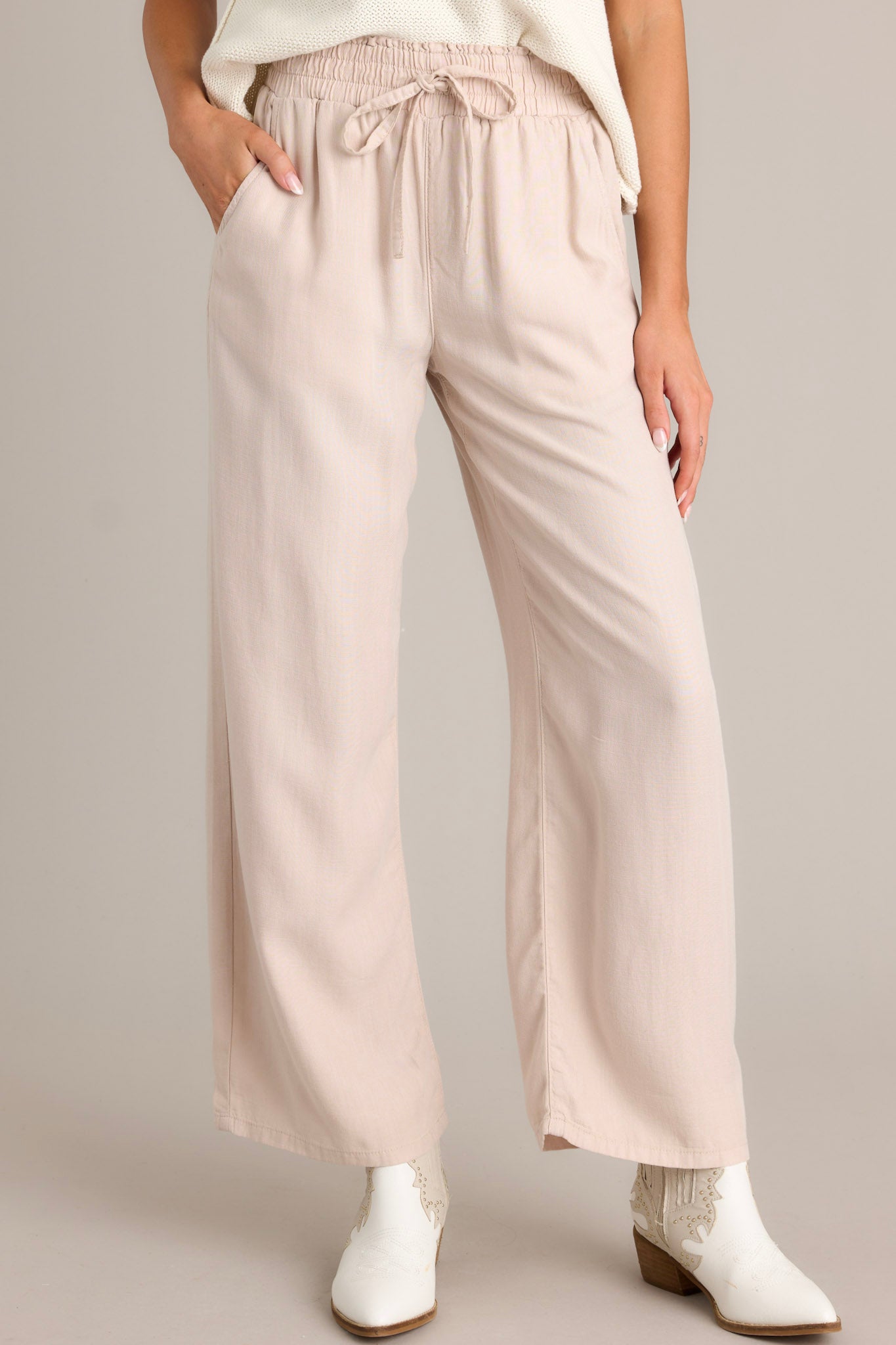 Front view of beige pants featuring a high waisted design, an elastic waistband with a self-tie drawstring, functional front & back pockets, and a wide leg.