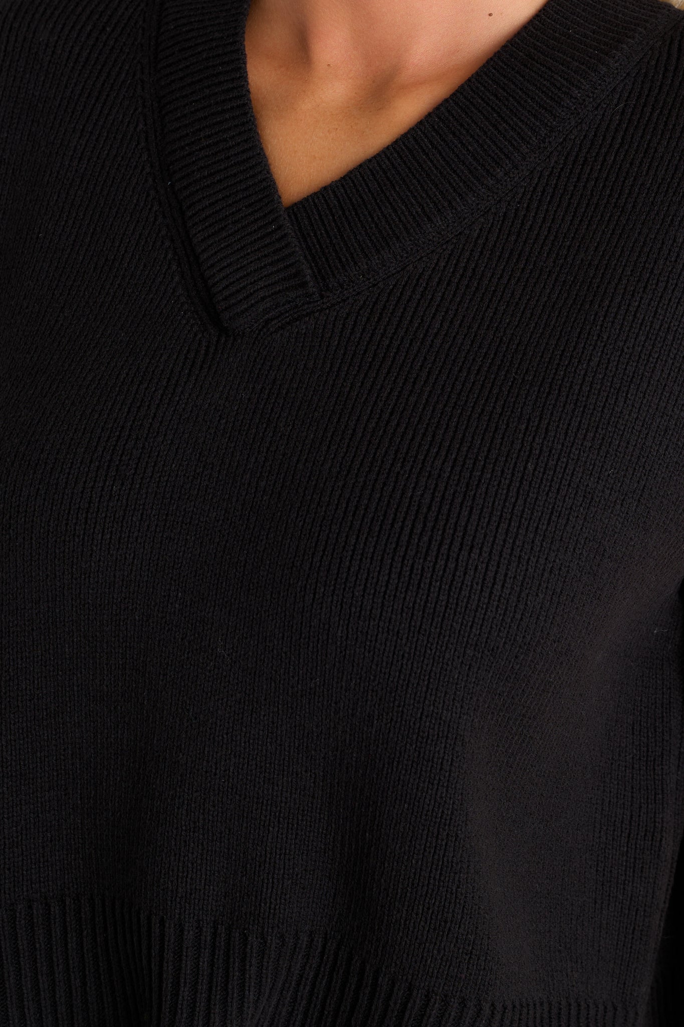 Close-up of the black sweater top showing the v-neckline, knit material, thick hemline, and side slits in the hem.