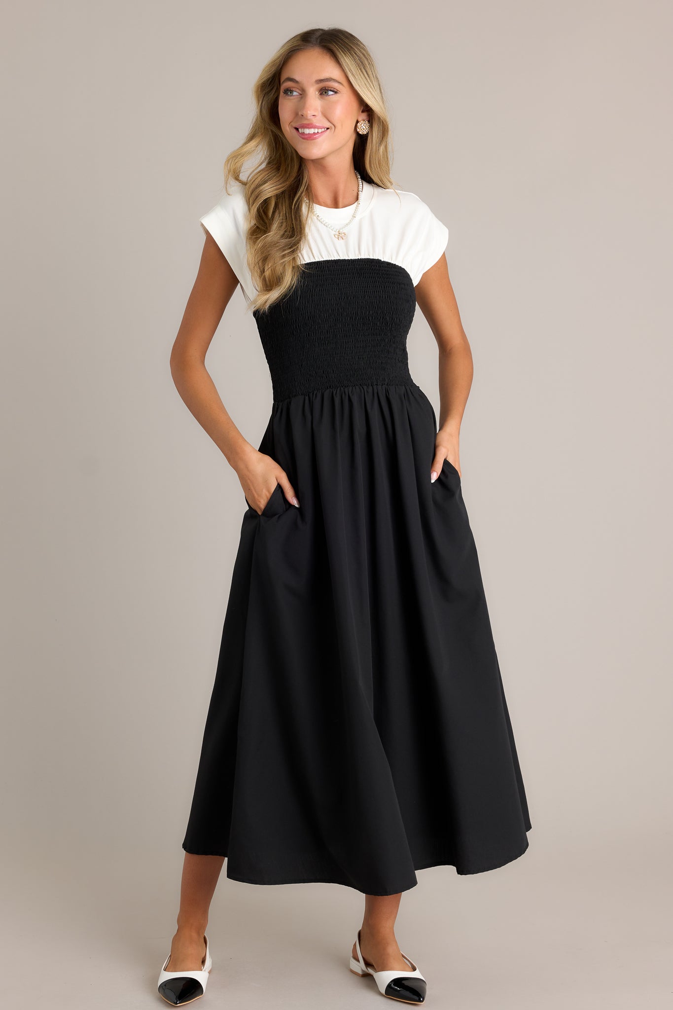 Front angled view of a black and white dress featuring a round neckline, a smocked waist, functional hip pockets, a white color-blocked top, and a flowing silhouette