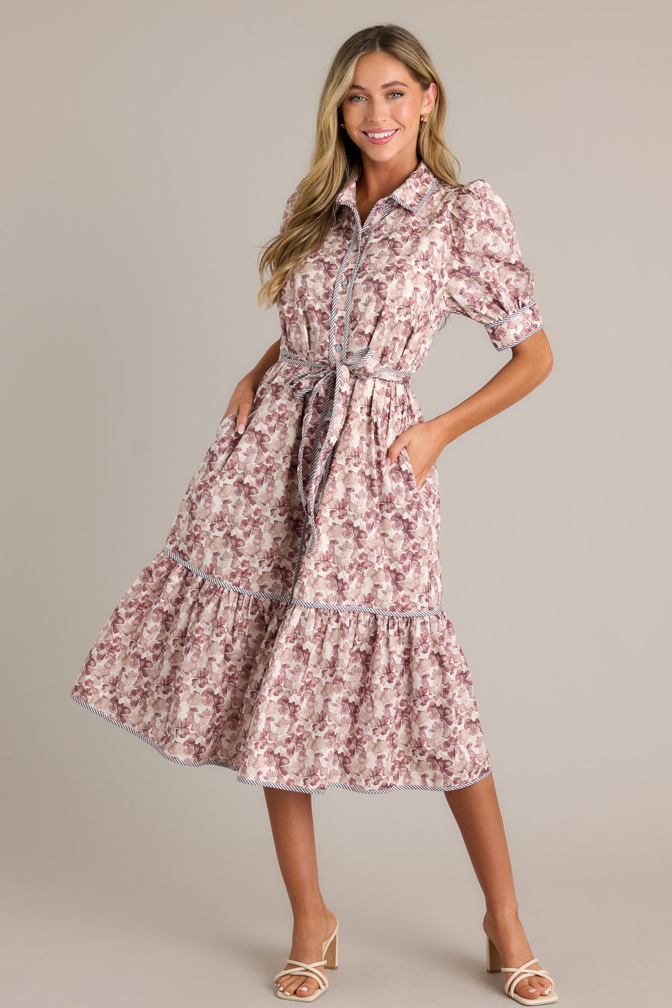 Action shot of a floral midi dress displaying the fit and movement, highlighting the collared neckline, functional button front, functional hip pockets, self-tie belt on the waist, tiered design, and puff sleeves.