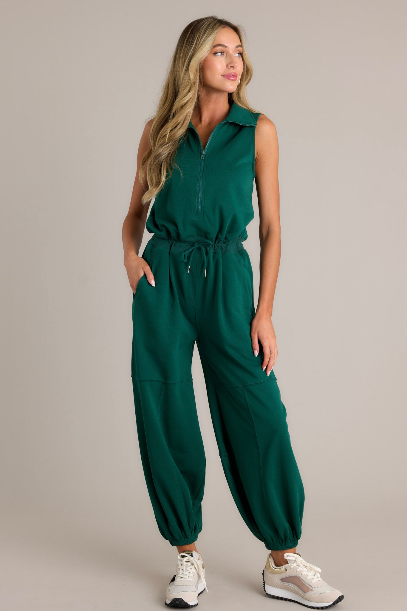 Full length view of a green jumpsuit with a collared neckline, a functional zipper front, a self-tie drawstring waist, functional hip pockets, elastic cuffed ankles, and a sleeveless design