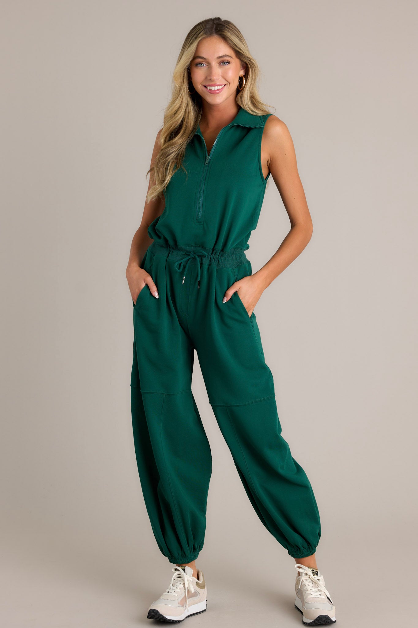 This green jumpsuit features a collared neckline, a functional zipper front, a self-tie drawstring waist, functional hip pockets, elastic cuffed ankles, and a sleeveless design.