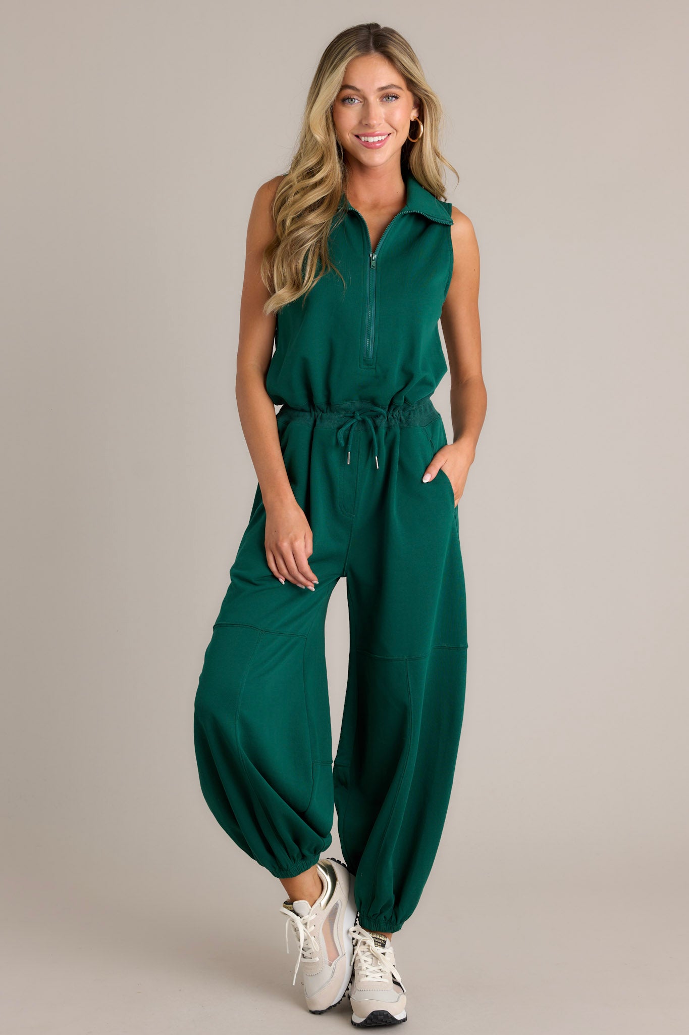 Front view of a green jumpsuit featuring a collared neckline, a functional zipper front, a self-tie drawstring waist, functional hip pockets, elastic cuffed ankles, and a sleeveless design