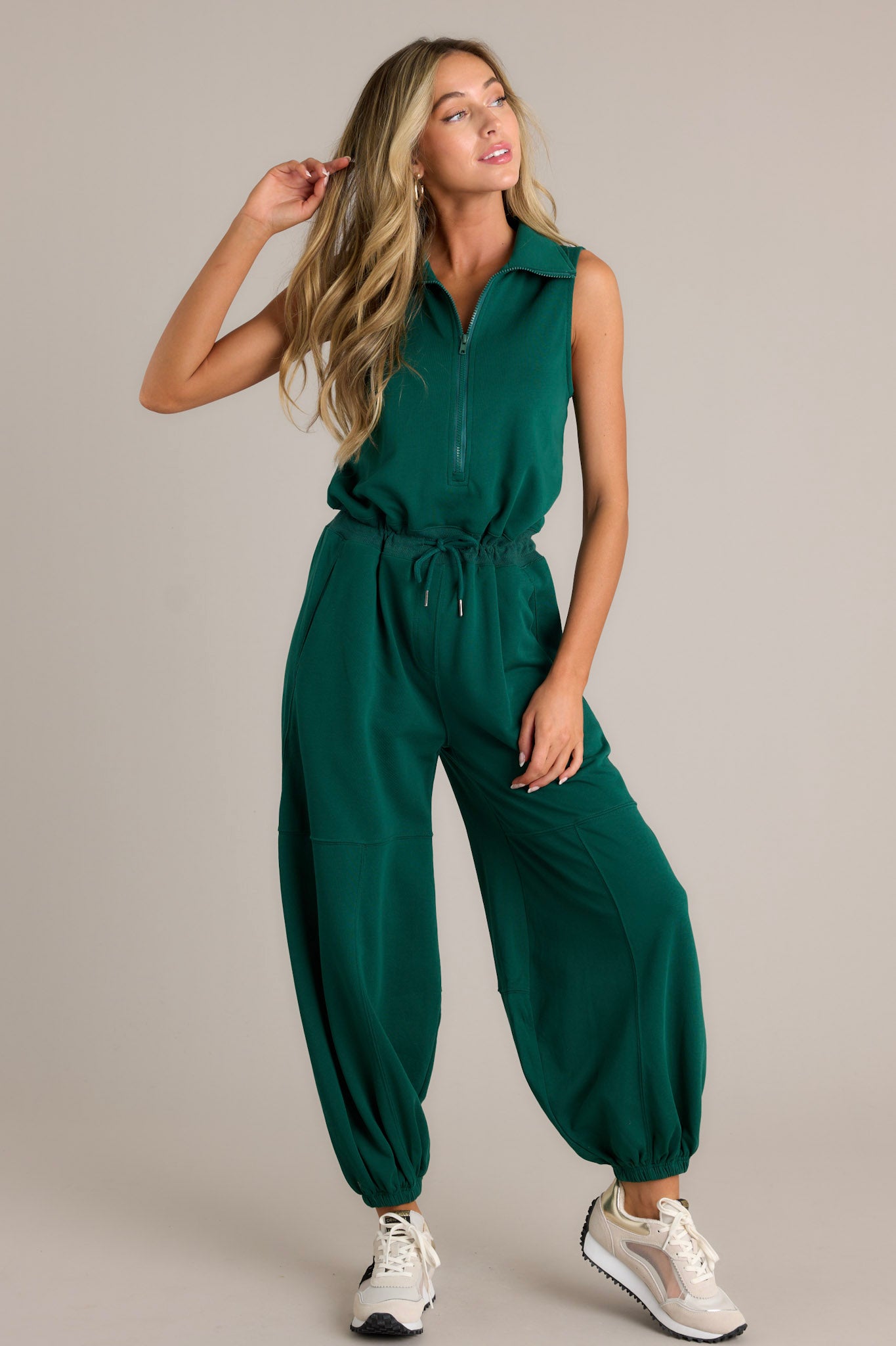 Front angled view of a green jumpsuit featuring a collared neckline, a functional zipper front, a self-tie drawstring waist, functional hip pockets, elastic cuffed ankles, and a sleeveless design