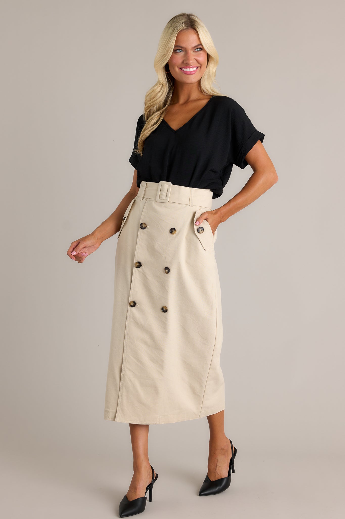 Full body view of this beige maxi skirt featuring a high waisted design, an elastic waist insert, a square fabric belt, functional hip pockets, functional front buttons and an open leg.