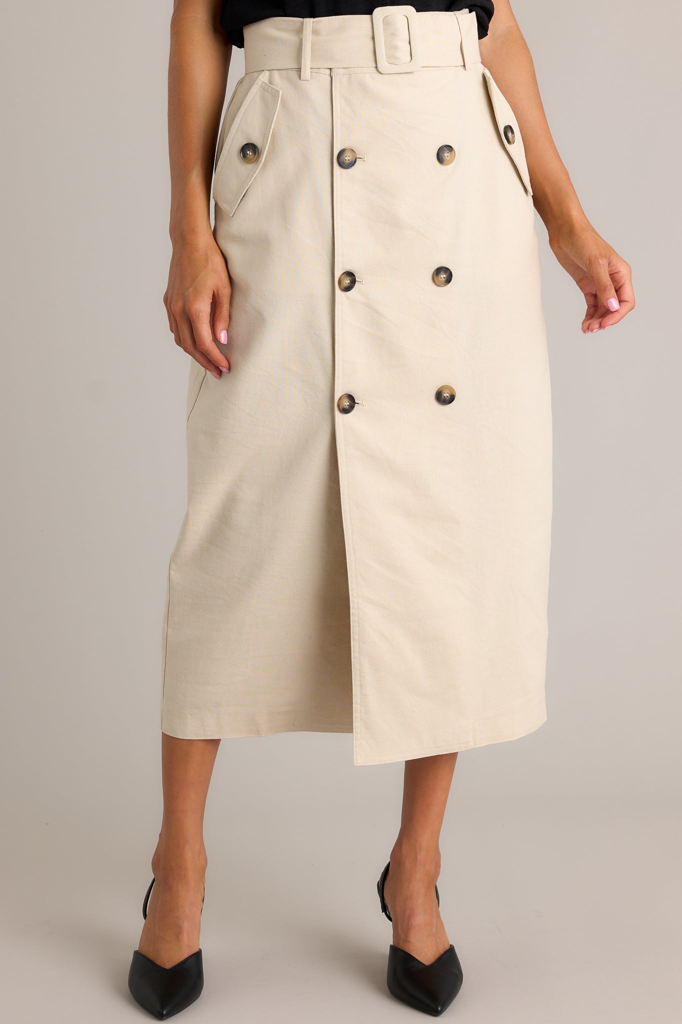 Front view of this beige maxi skirt featuring a high waisted design, an elastic waist insert, a square fabric belt, functional hip pockets, functional front buttons and an open leg.