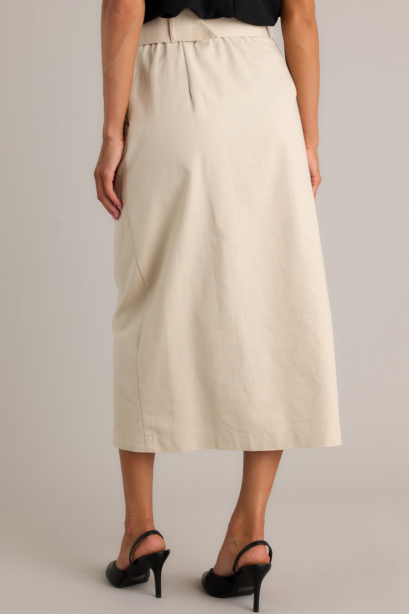 Back view of this beige maxi skirt featuring a high waisted design, an elastic waist insert, a square fabric belt, functional hip pockets, functional front buttons and an open leg.