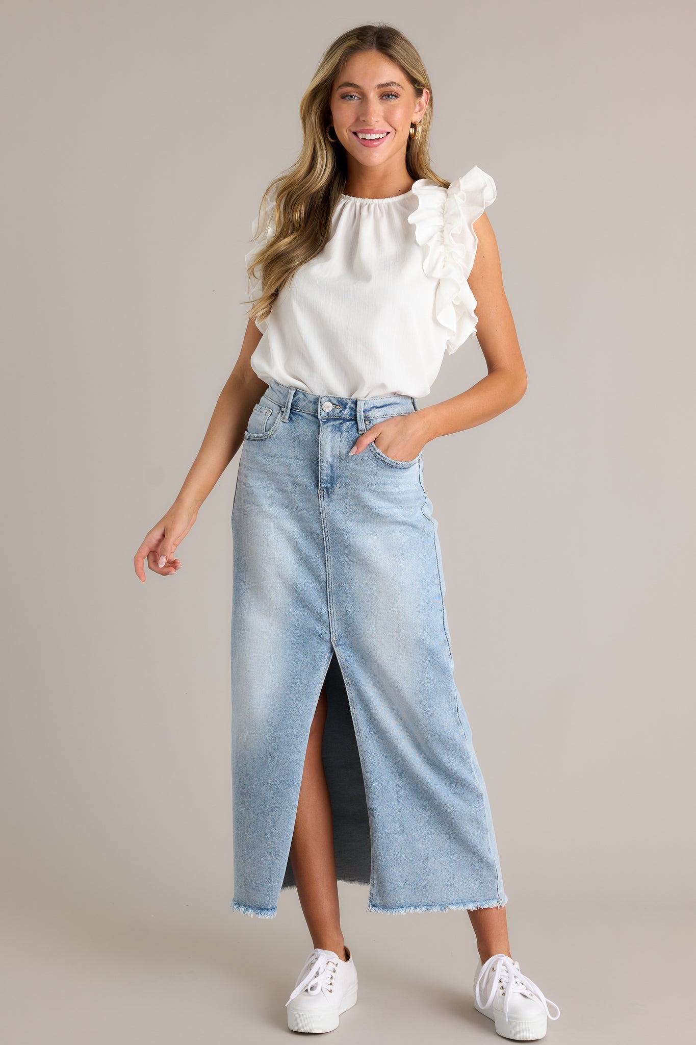 This light wash denim midi skirt features a high waisted design, belt loops, a zipper and button closure, functional pockets, a 15" slit up the center, and raw hem detailing.