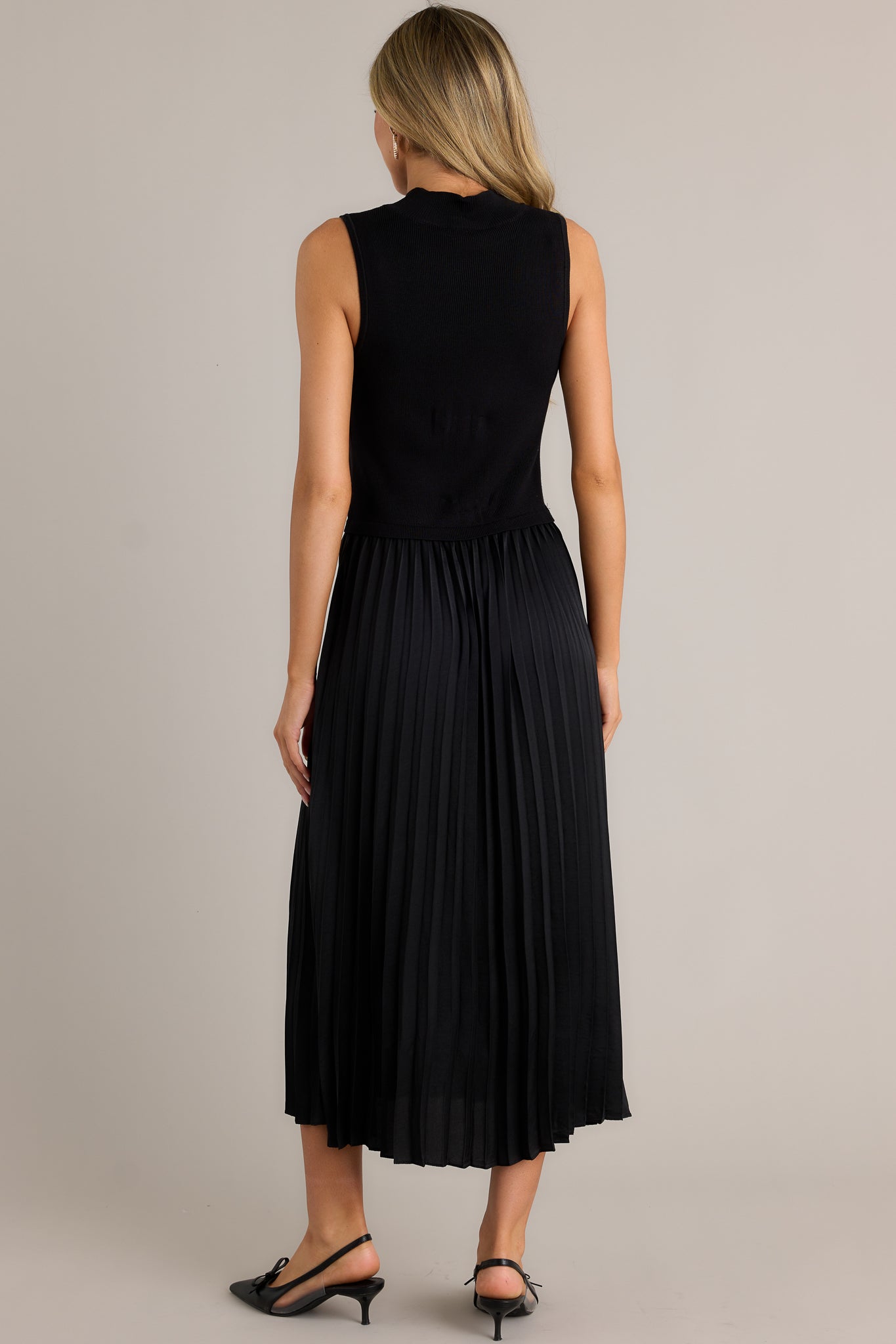 Back view of this black sleeveless dress featuring a high neckline, a ribbed knit bodice, a pleated skirt, a sleeveless design and a flowing silhouette.