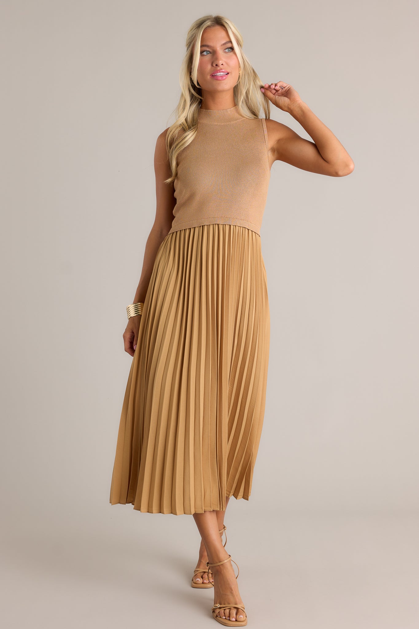 Posed view of this mustard sleeveless dress featuring a high neckline, a ribbed knit bodice, a pleated skirt, a sleeveless design and a flowing silhouette.