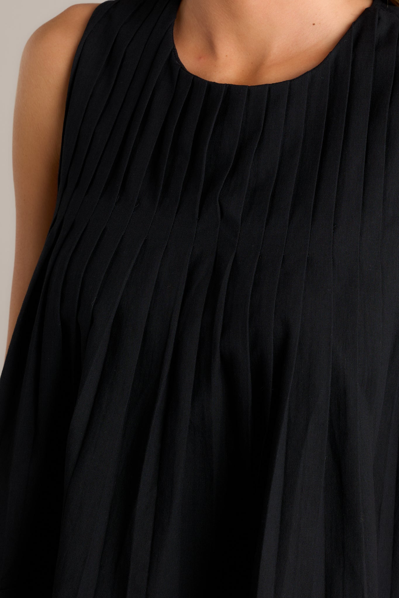 Close-up of the sleeveless black top focusing on the round neckline, pleated bust, and the texture of the slightly pleated bodice.