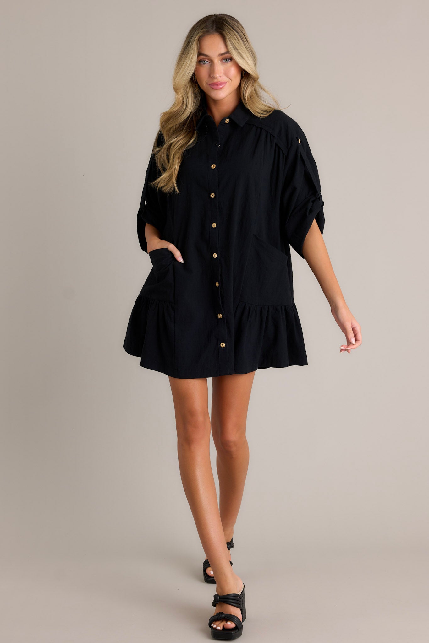 A black dress featuring a collared high neckline, functional button front, and large front pockets. The dress has loose sleeves and a ruffled hem.