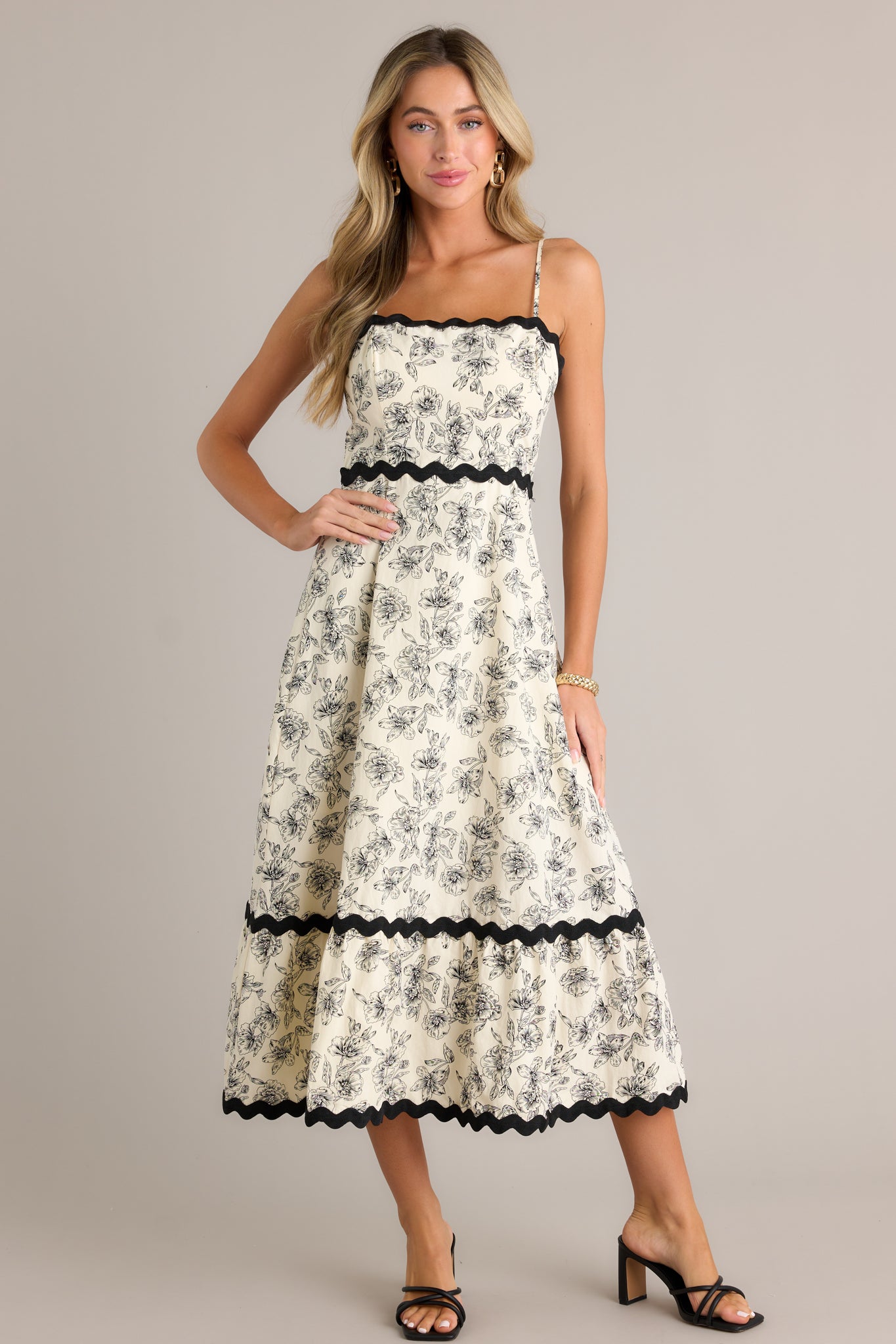 Front view of an ivory maxi dress featuring a square neckline, black ruffle trim, a tiered design, a black floral print, a discrete back zipper, and adjustable spaghetti straps.