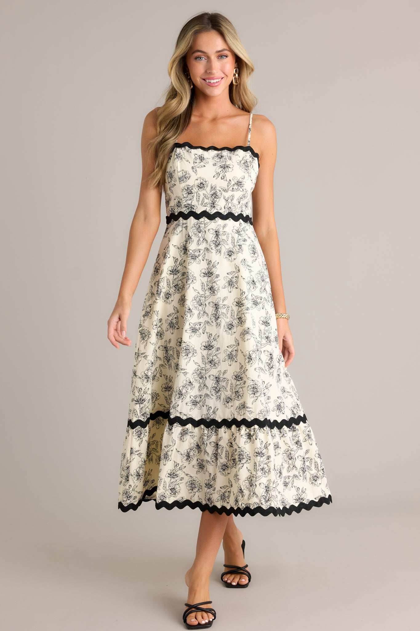 Full length view of an ivory maxi dress featuring a square neckline, black ruffle trim, a tiered design, a black floral print, a discrete back zipper, and adjustable spaghetti straps