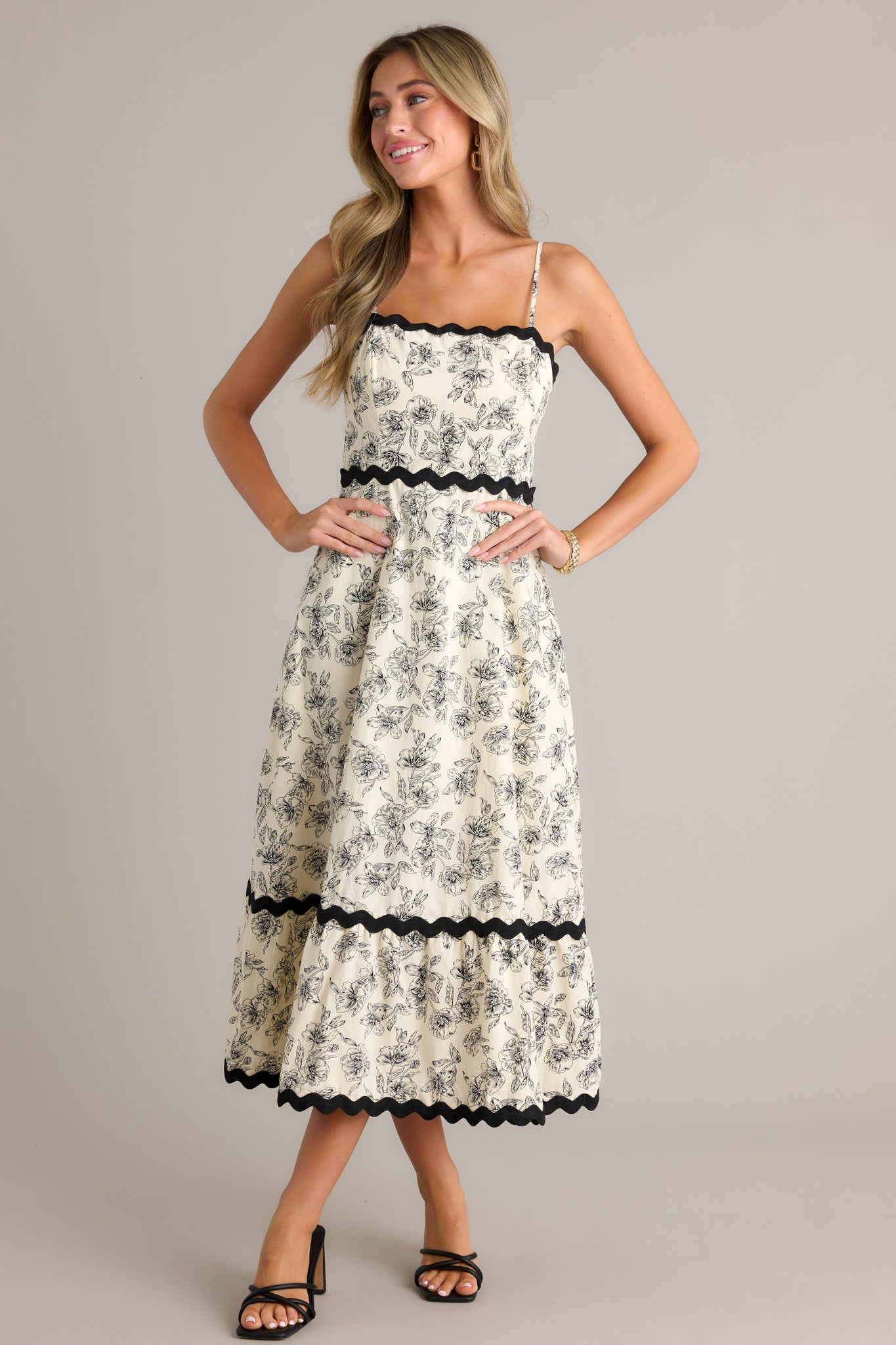 This ivory maxi dress features a square neck, black ruffle trim, a tiered design, a black floral print, a discrete back zipper and adjustable spaghetti straps.