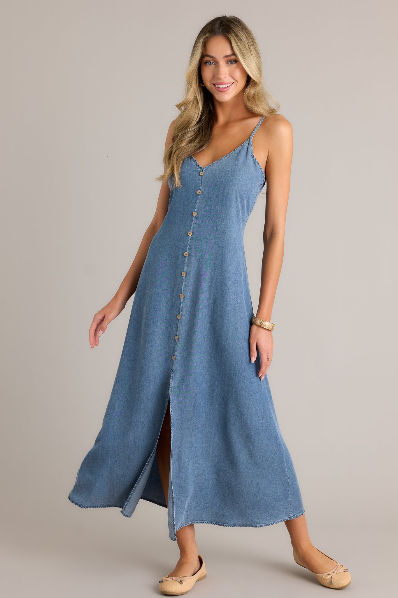 Action shot of a chambray maxi dress displaying the fit and movement, highlighting the v-neckline, button front design, smocked back insert, adjustable spaghetti straps, and open leg slit.