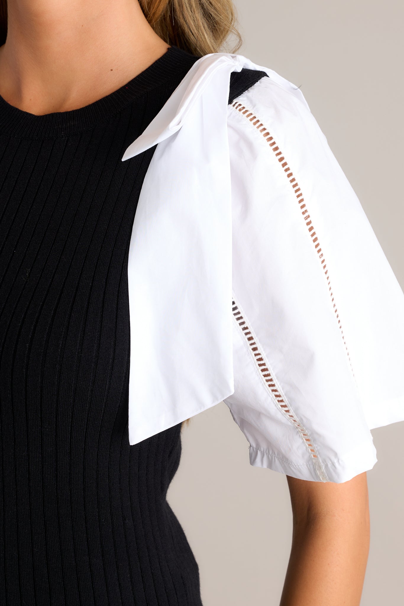 Close-up of the black and white sweater top showing the round neckline, ribbed sweater bodice, bowties on each shoulder, and delicate cut-out detailing on the sleeves.