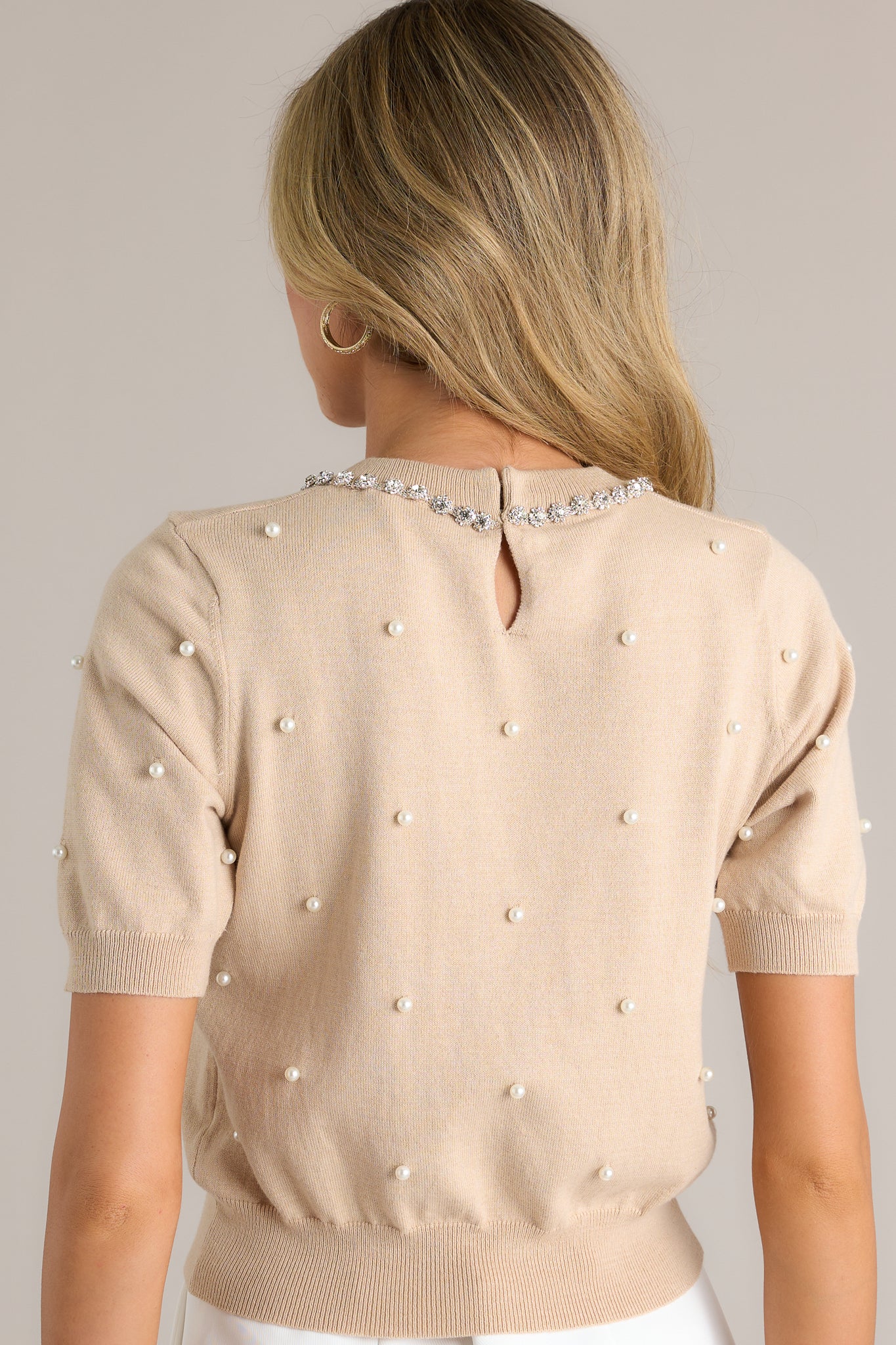 Back view of this beige short sleeve top featuring a rounded neckline, a rhinestone chained collar, faux pearls, a thick hemline and slightly cuffed short sleeves.