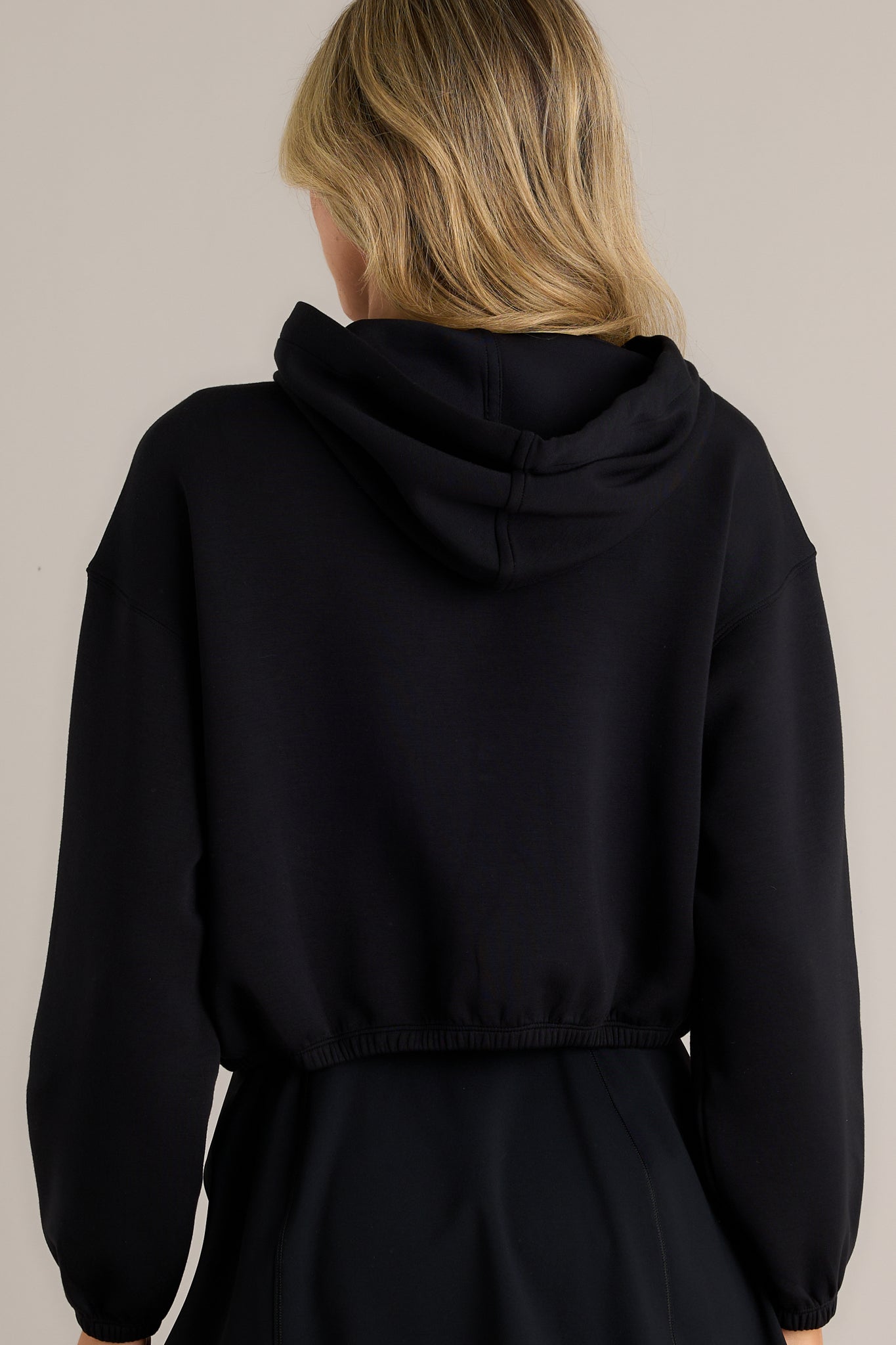 Back view of a black hoodie highlighting the overall fit, slightly cropped design, elastic hem, and elastic cuffed sleeves.