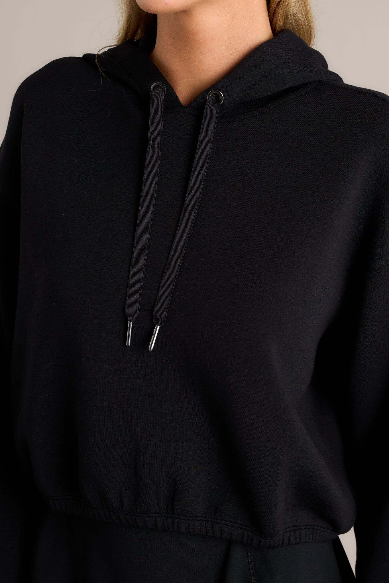 Close-up of the black hoodie showing the high neckline, draw cord, elastic hem, and finely knit fabric.