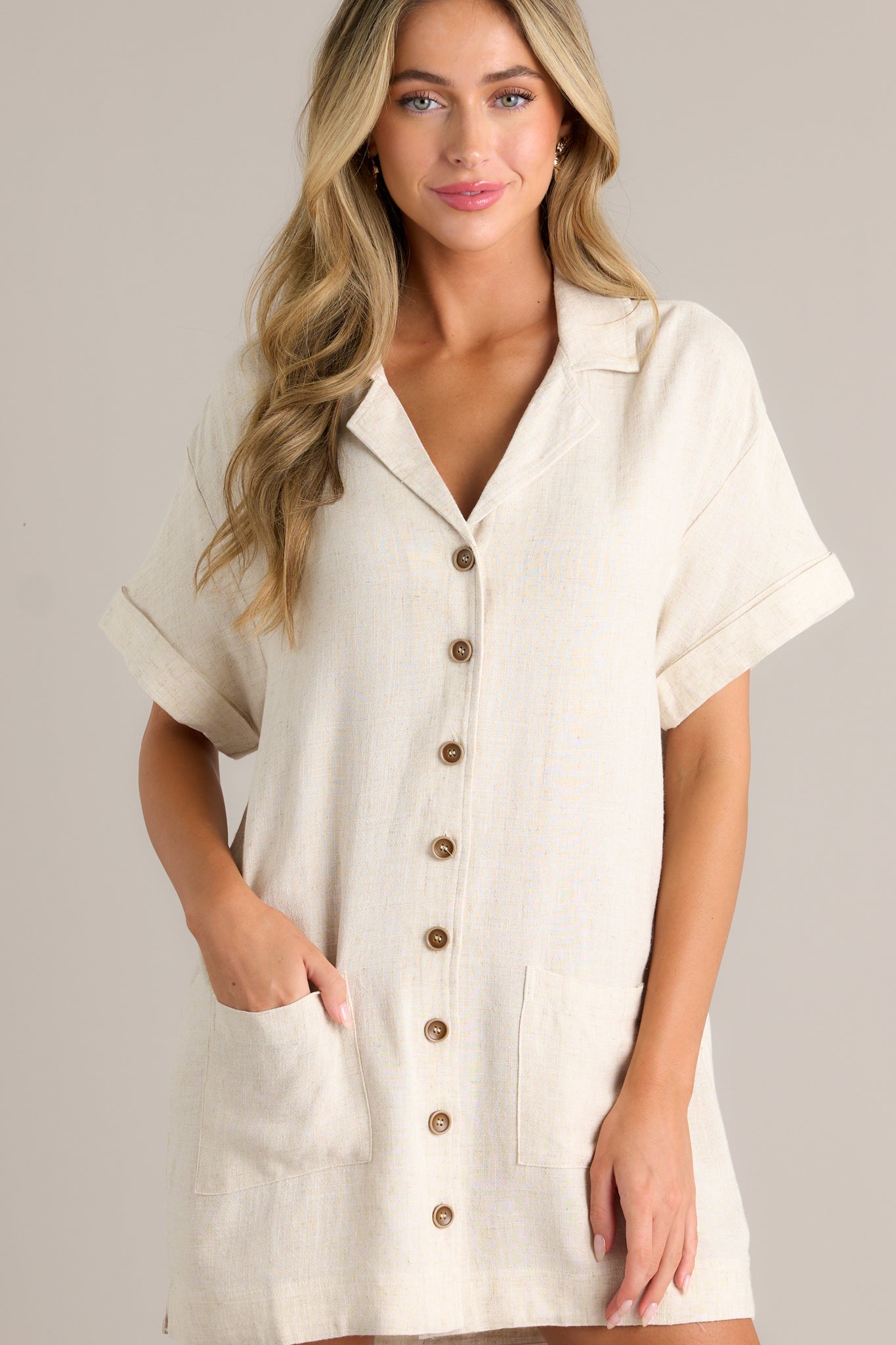 Front view of a beige dress featuring a collared v-neckline, functional button front, functional hip pockets, short cuffed sleeves, and a relaxed fit.