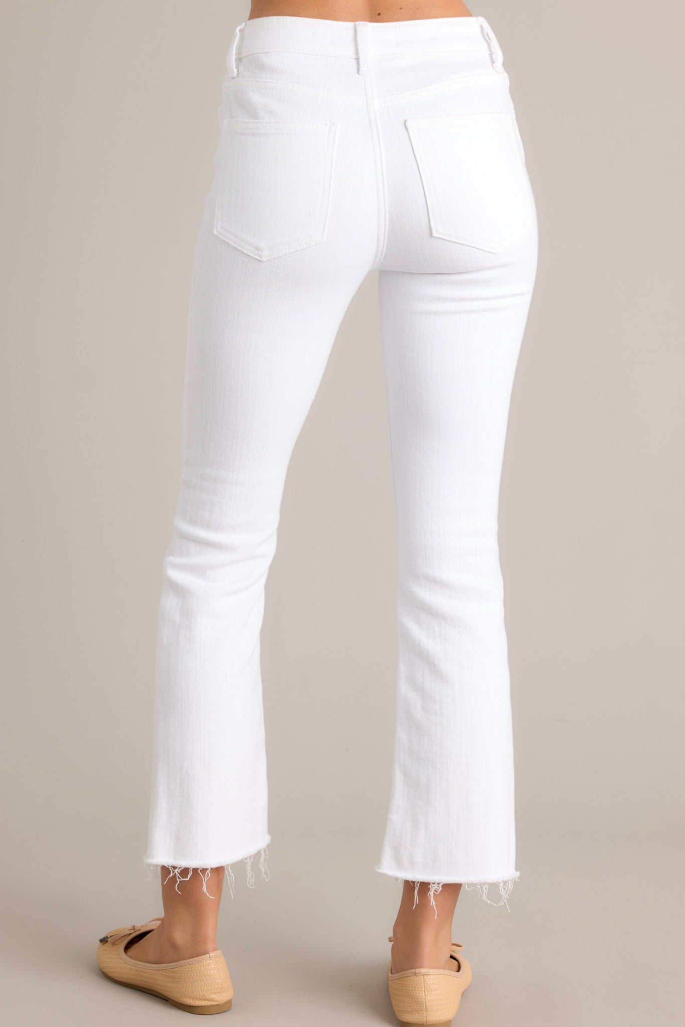 Back view of these white jeans that feature a high waisted design, a classic button & zipper closure, belt loops, functional front & back pockets, and a raw, cropped hemline.