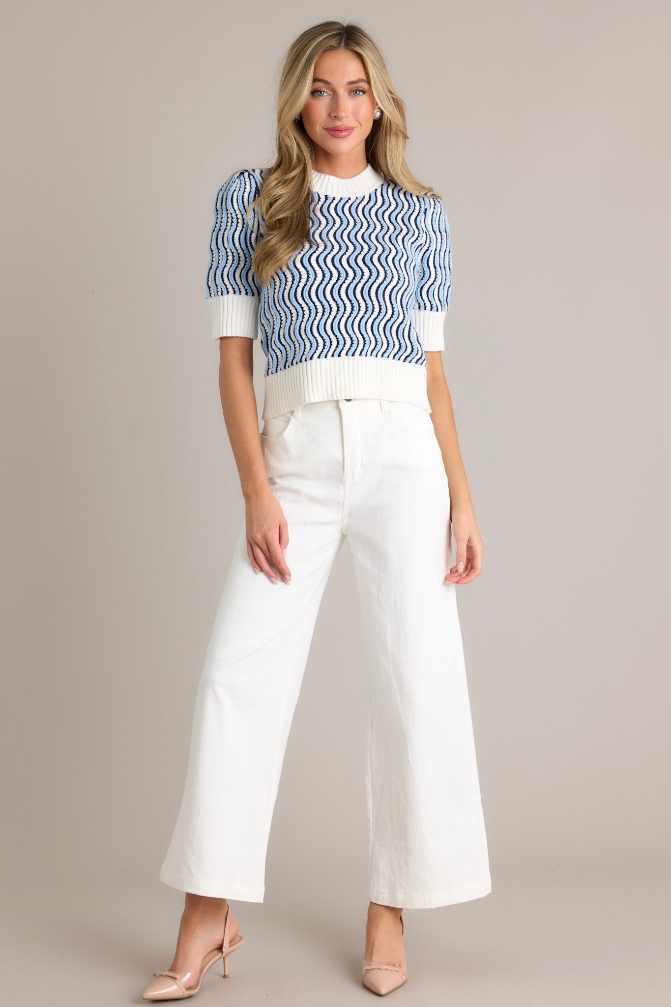 Full length view of a model wearing a blue stripe sweater top with a high knit neckline, wavy blue and white stripe design, thick hemline, ivory color blocking, short sleeves, and a soft knit fabric.