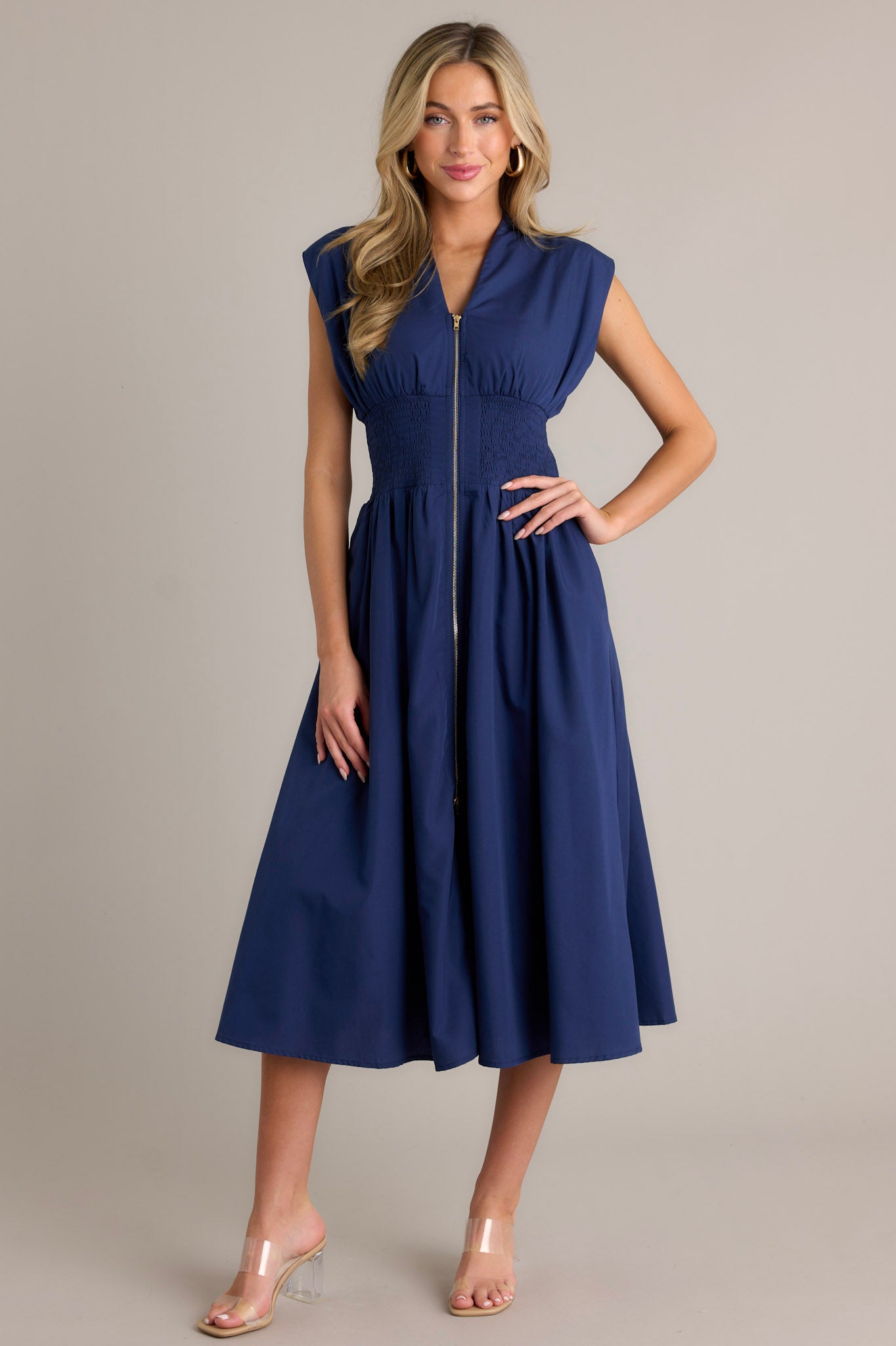 Full body view of a navy blue dress with a V-neckline, cap sleeves, a zippered front, and a cinched waist with shirred detailing, featuring a flowing skirt and side pockets.