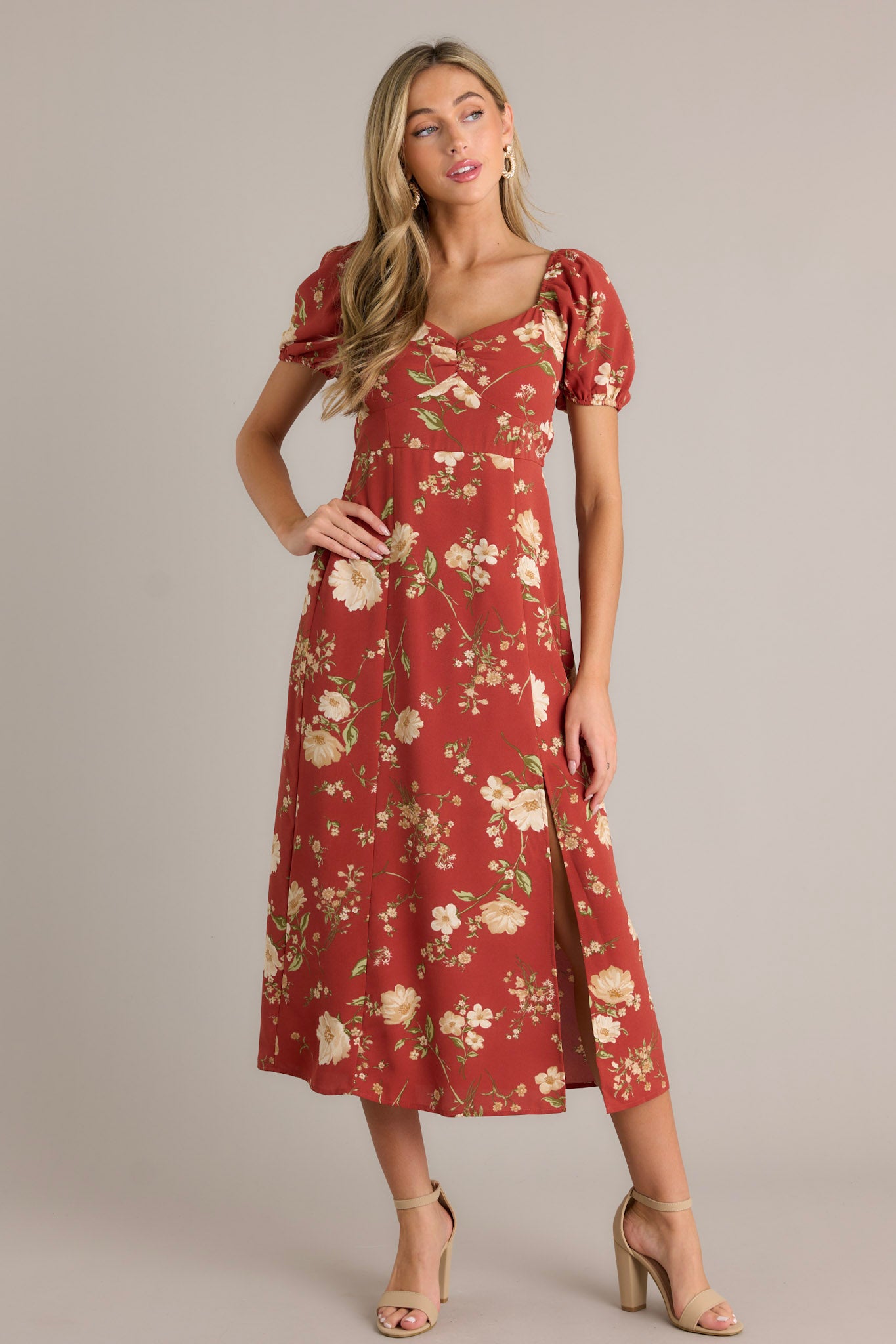 Full body view of this burnt orange dress that features a square neckline with a slight v-neckline, a floral design, a smocked back insert, a slit in the left leg and short cinched puff sleeves.