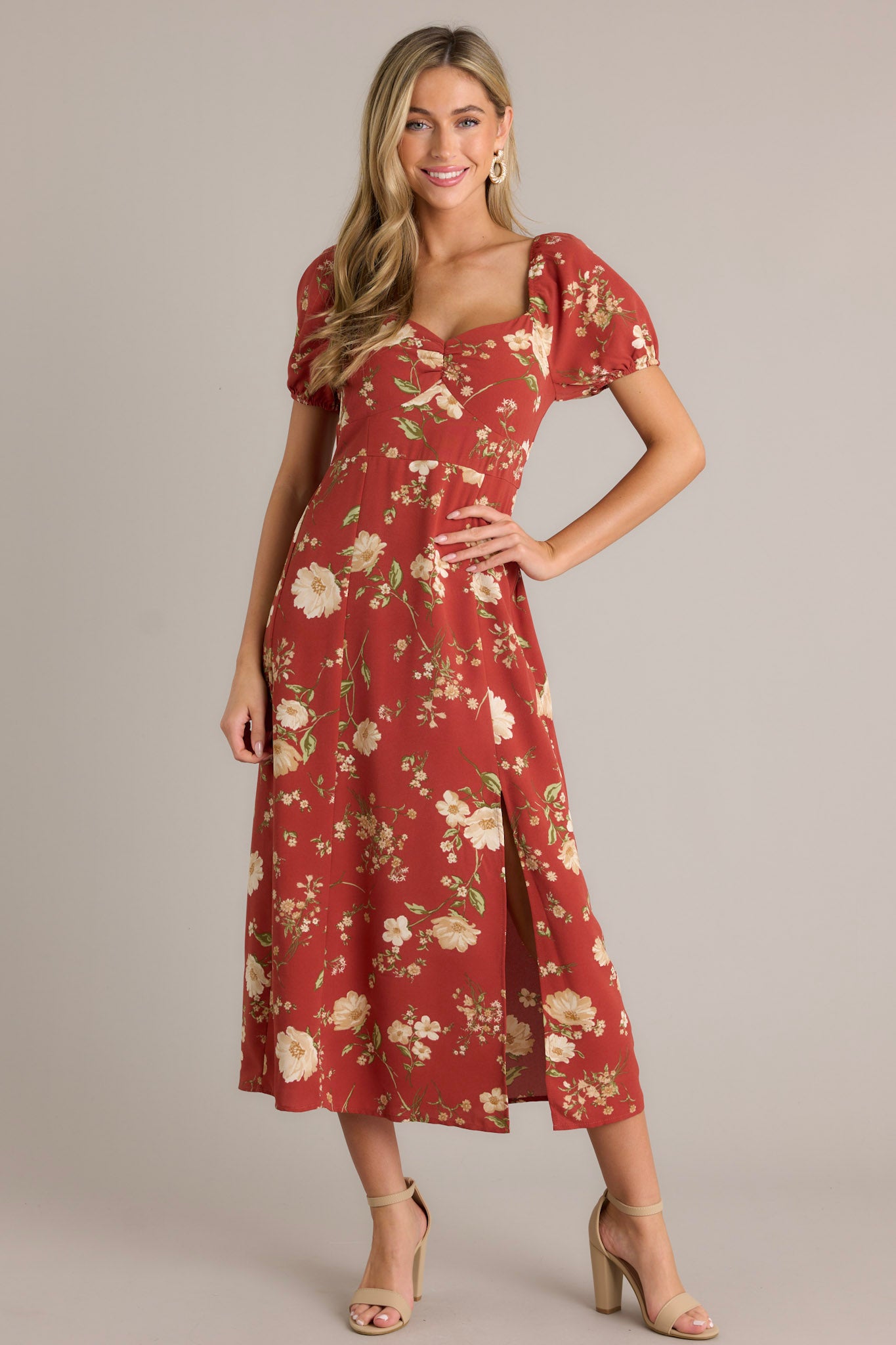 Front angled view of this burnt orange dress that features a square neckline with a slight v-neckline, a floral design, a smocked back insert, a slit in the left leg and short cinched puff sleeves.