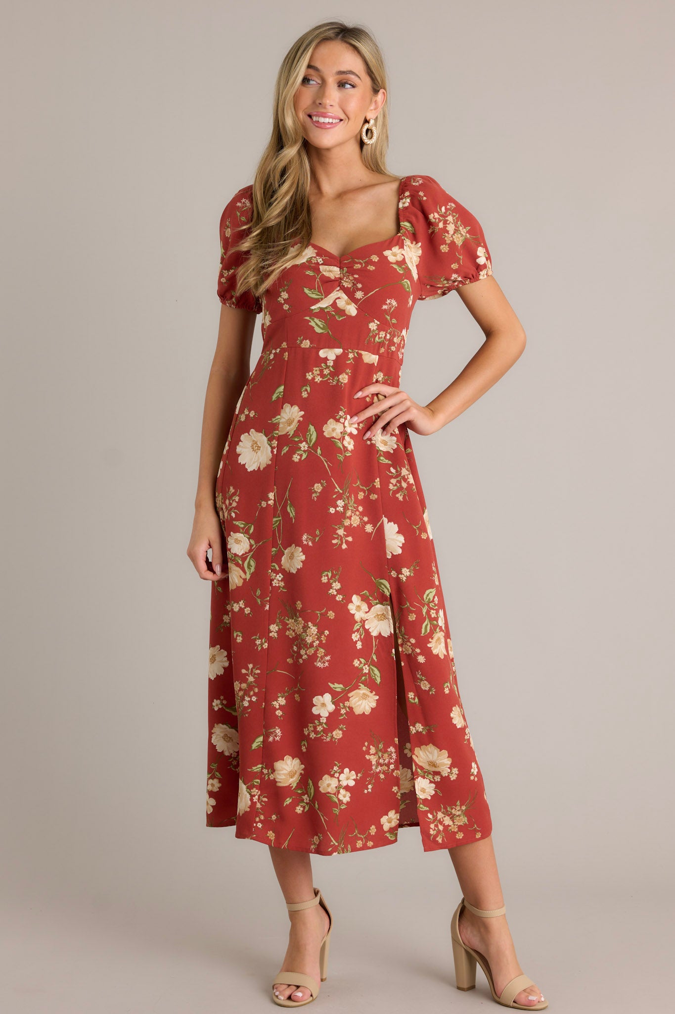 Full length view of this burnt orange dress that features a square neckline with a slight v-neckline, a floral design, a smocked back insert, a slit in the left leg and short cinched puff sleeves.