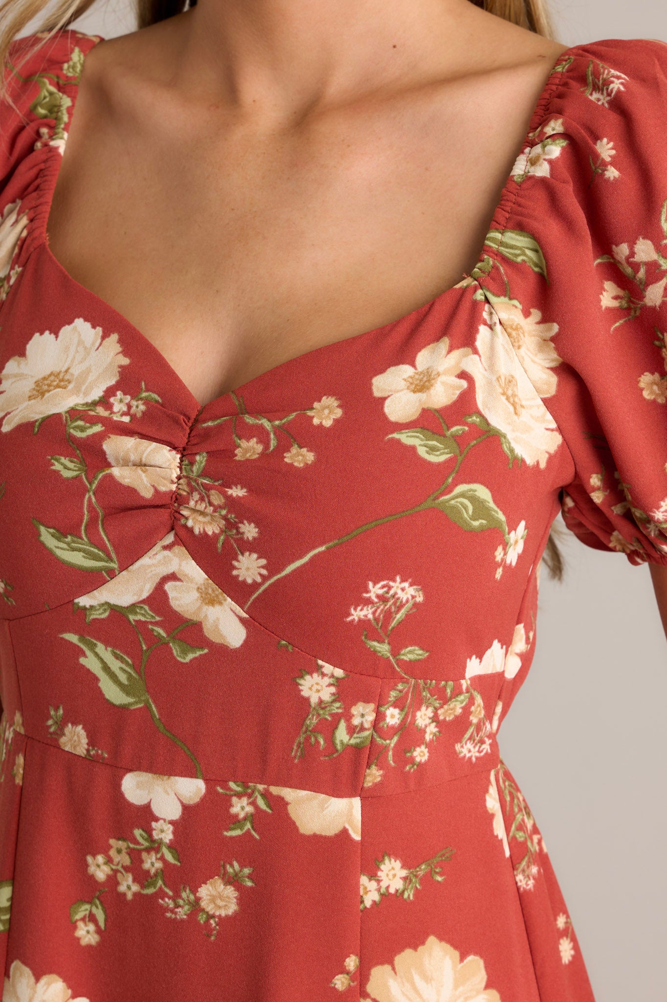 Close-up view of this burnt orange dress that features a square neckline with a slight v-neckline, a floral design, a smocked back insert, a slit in the left leg and short cinched puff sleeves.