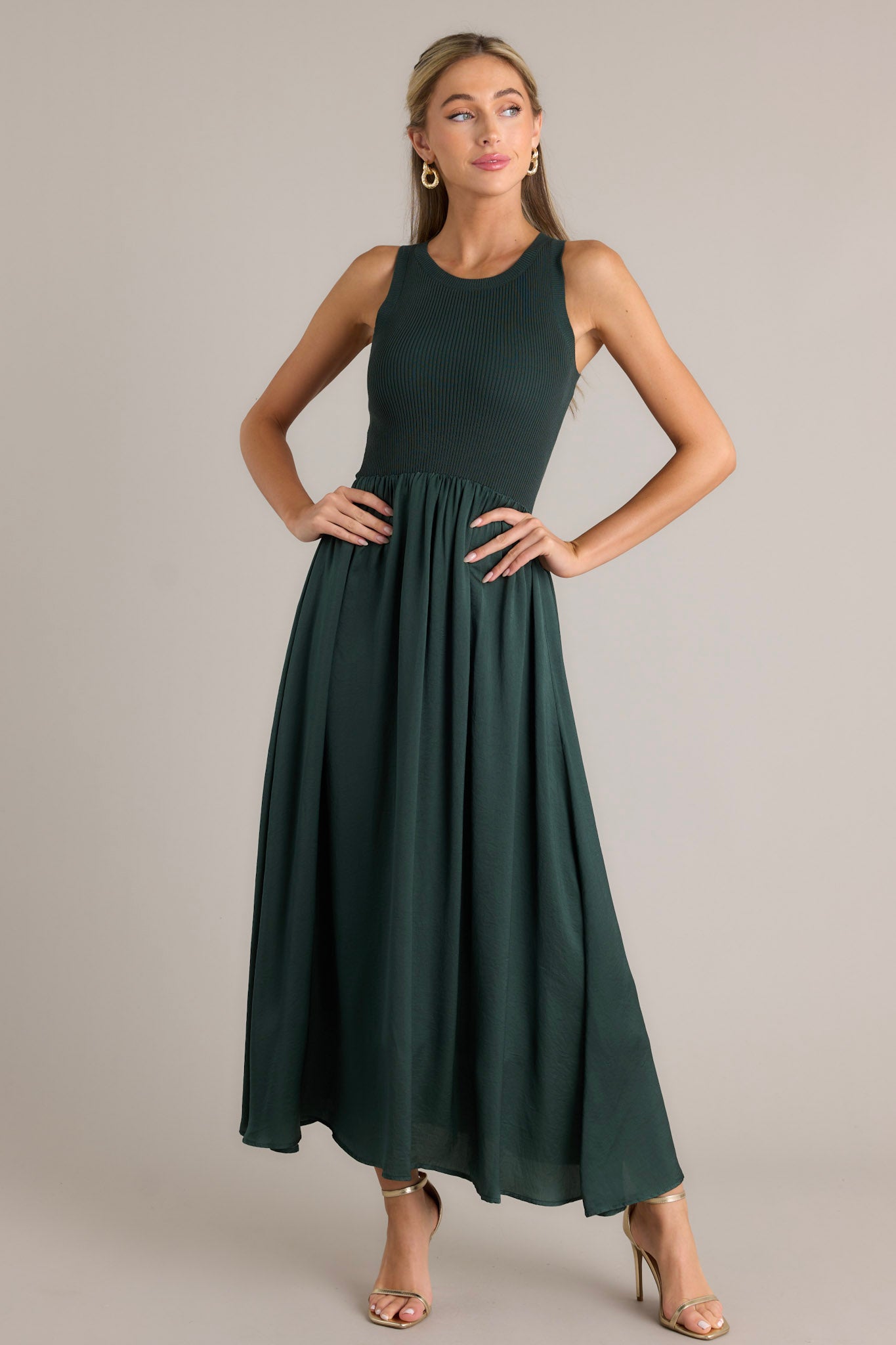 Front angled view of a sweater top dress featuring a high rounded neckline, ribbed sweater bodice, lightly pleated skirt, a sleeveless design, and a flowing silhouette