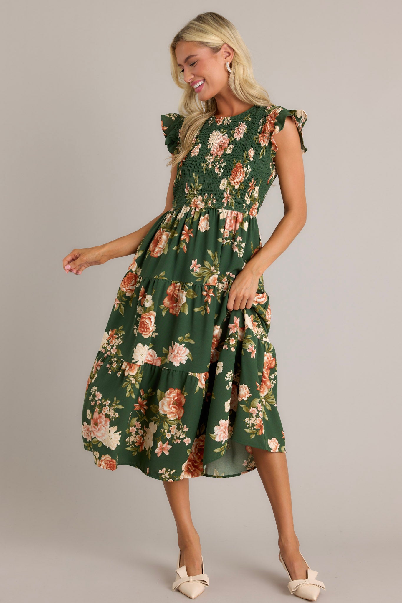 Front angled view of a green dress featuring a high neckline, a fully smocked bodice, a tiered design, short ruffle sleeves, a back button and loop closure, and a flowing silhouette