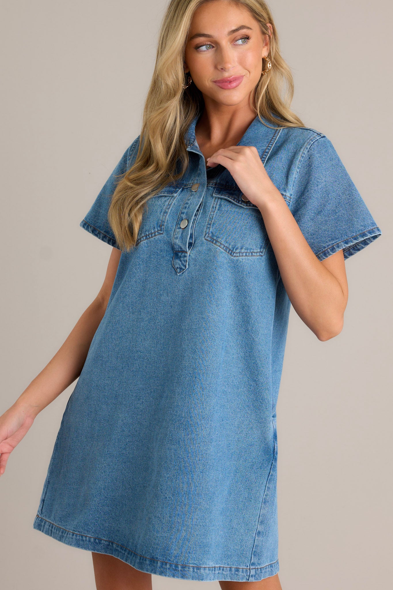 Front angled view of a denim mini dress featuring a collared neckline, functional button front bust, functional bust pockets with button closures, functional hip pockets, and short sleeves