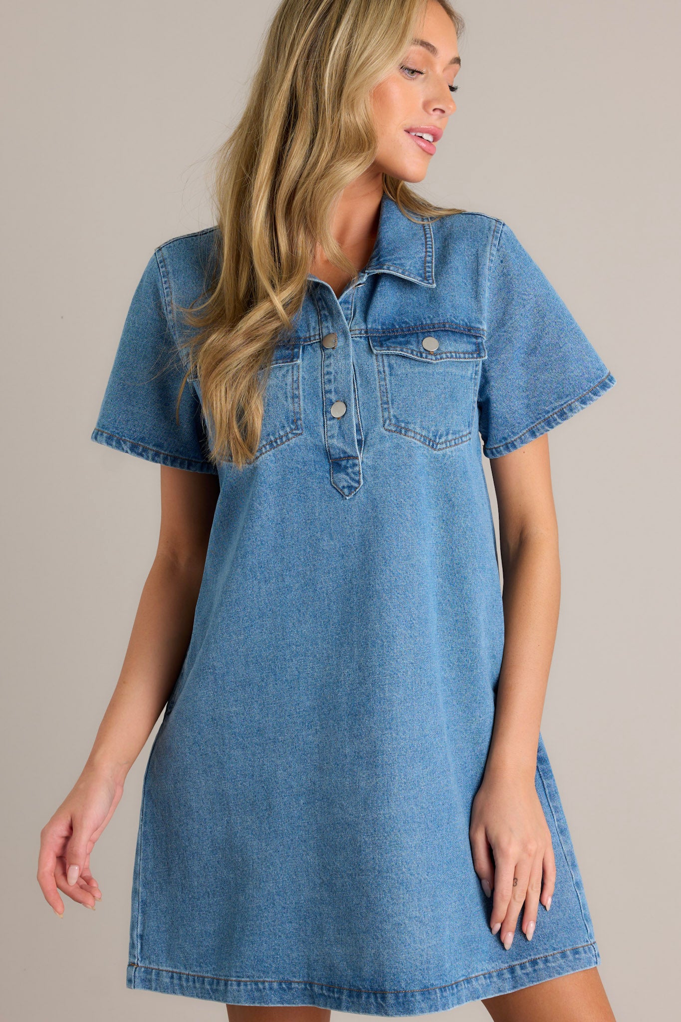 Front view of a denim mini dress featuring a collared neckline, a functional button front bust, functional bust pockets with button closures, functional hip pockets, and short sleeves.