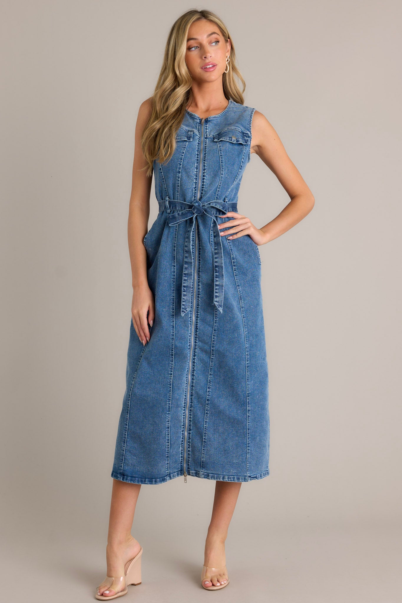 Front angled view of a denim dress featuring a rounded neckline, functional front zipper, faux chest pockets, functional hip pockets, a self-tie denim drawstring belt, and a sleeveless design,