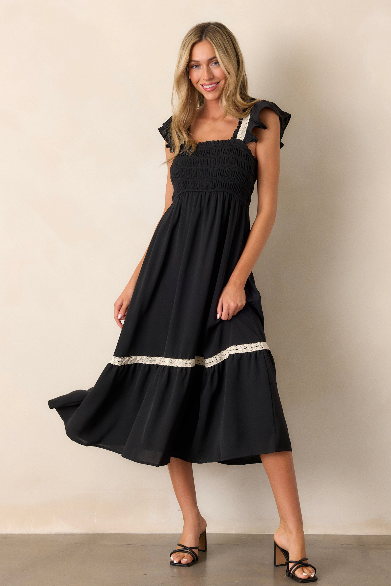 Full length view of a black dress featuring a smocked bodice, square neckline, tiered design, white lace detailing on the sleeves and trim, and short ruffle sleeves