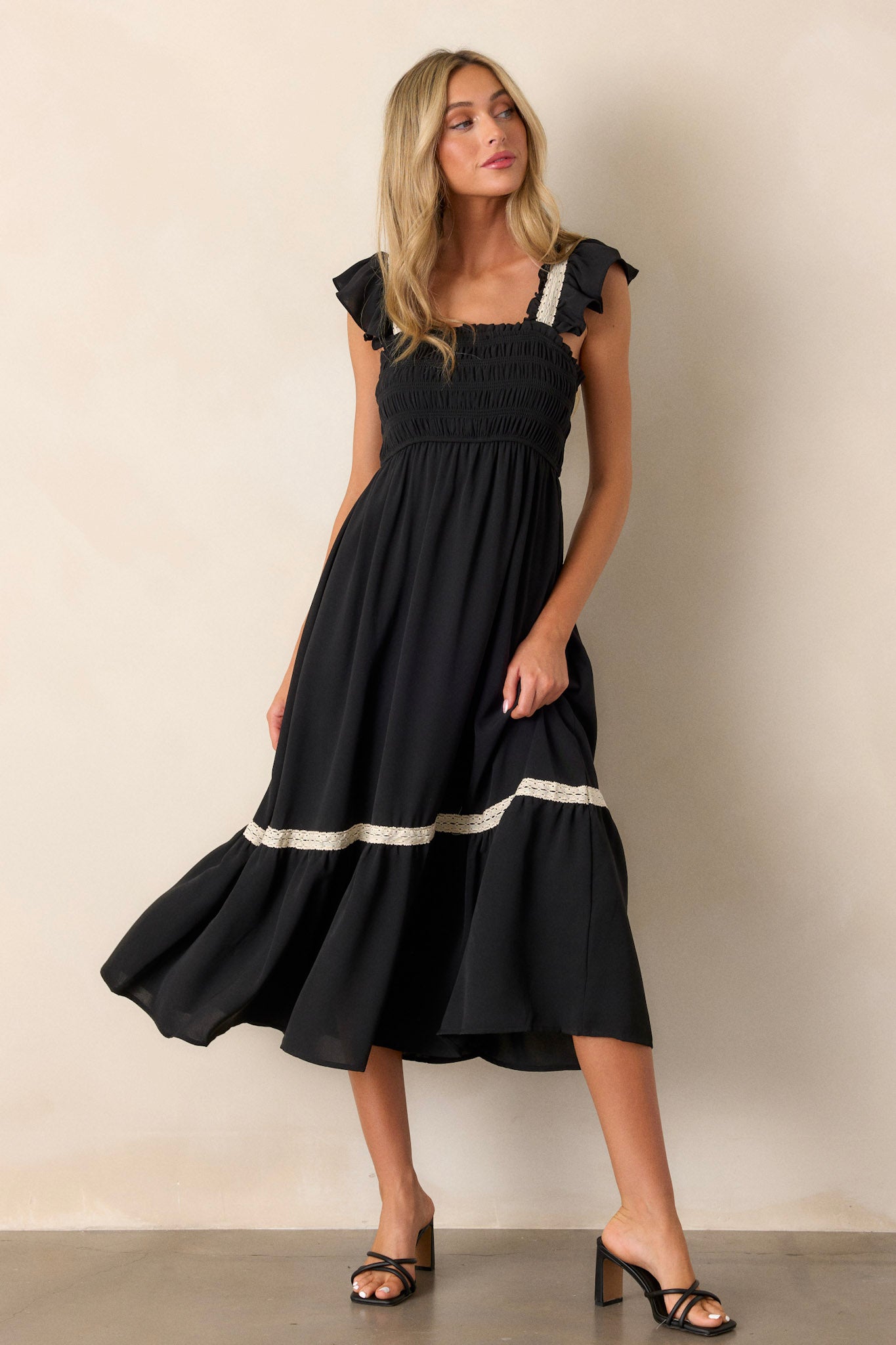 Front view of a black dress featuring a smocked bodice, a square neckline, a tiered design, white lace detailing on the sleeves, and trim on the tier with short ruffle sleeves.