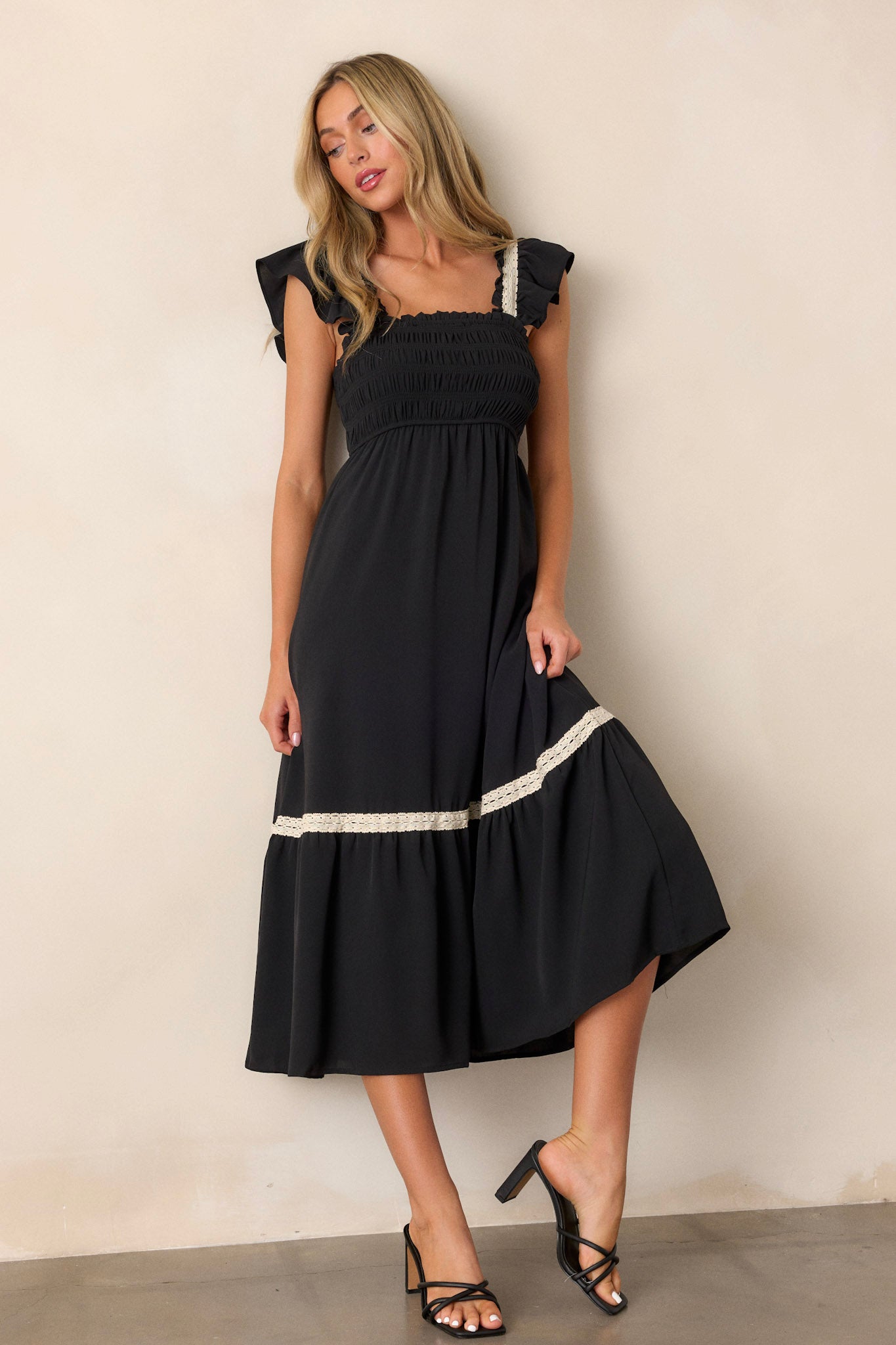 This black dress features a smocked bodice, a square neckline, a tiered design, white lace detailing on the sleeves and trim on the tier and short ruffle sleeves.