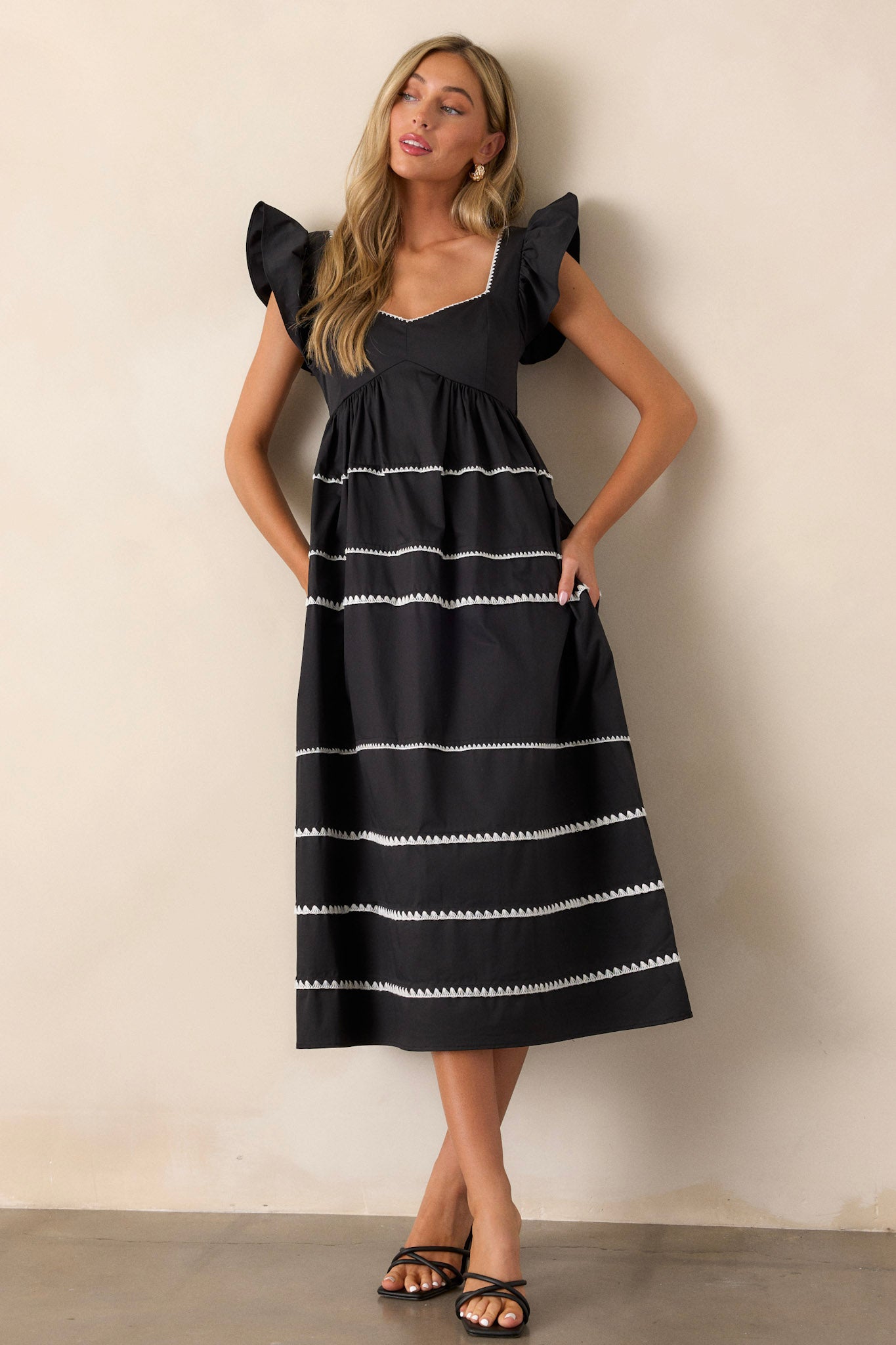 Full length view of a black dress featuring ruffle cap sleeves, a smocked back, multiple tiers, two functional pockets, and ric-rac detailing