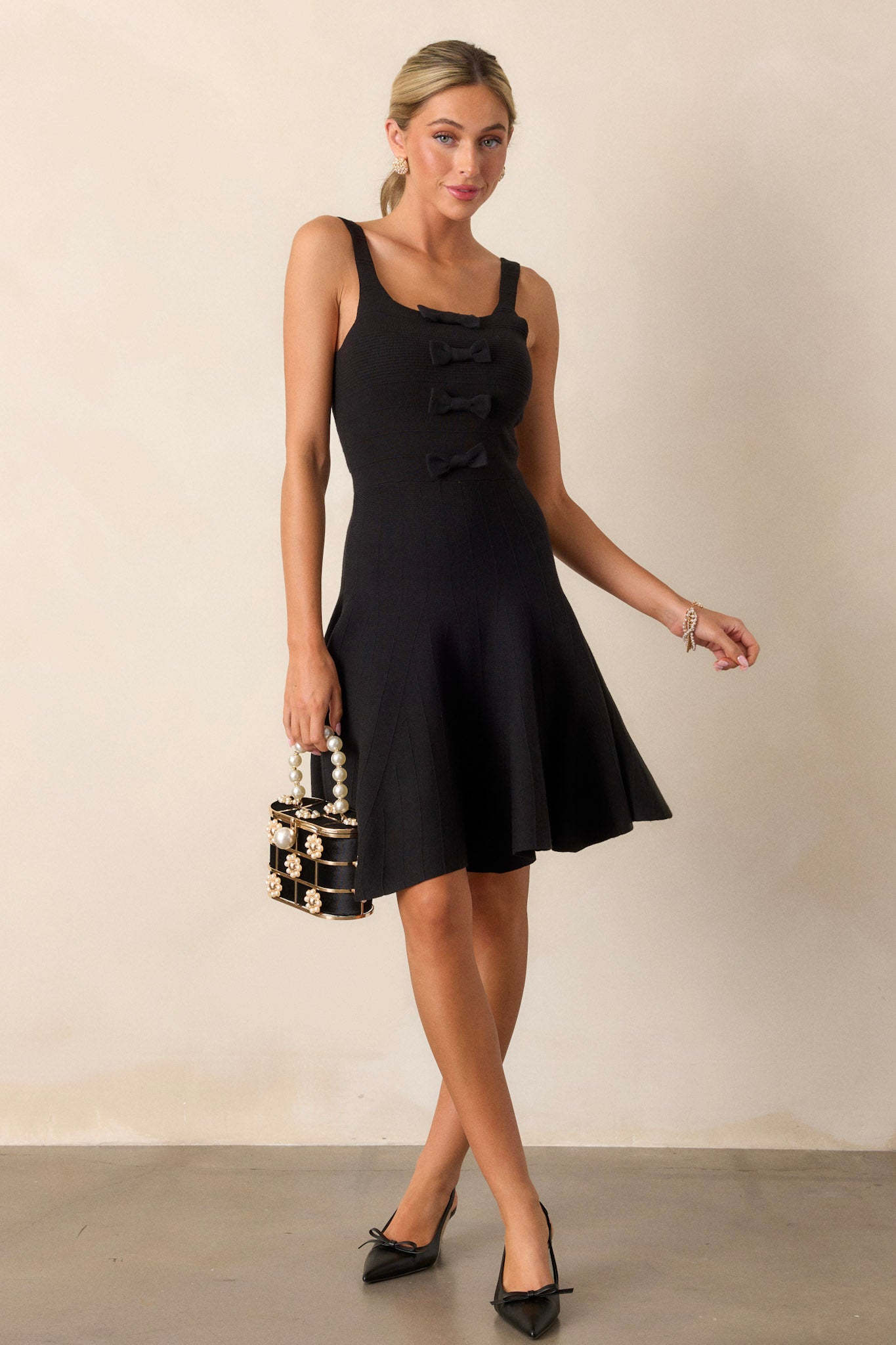 Action shot of the black sleeveless dress displaying the flow and movement of the slightly pleated skirt, emphasizing the square neckline, bow details, and soft knit texture.